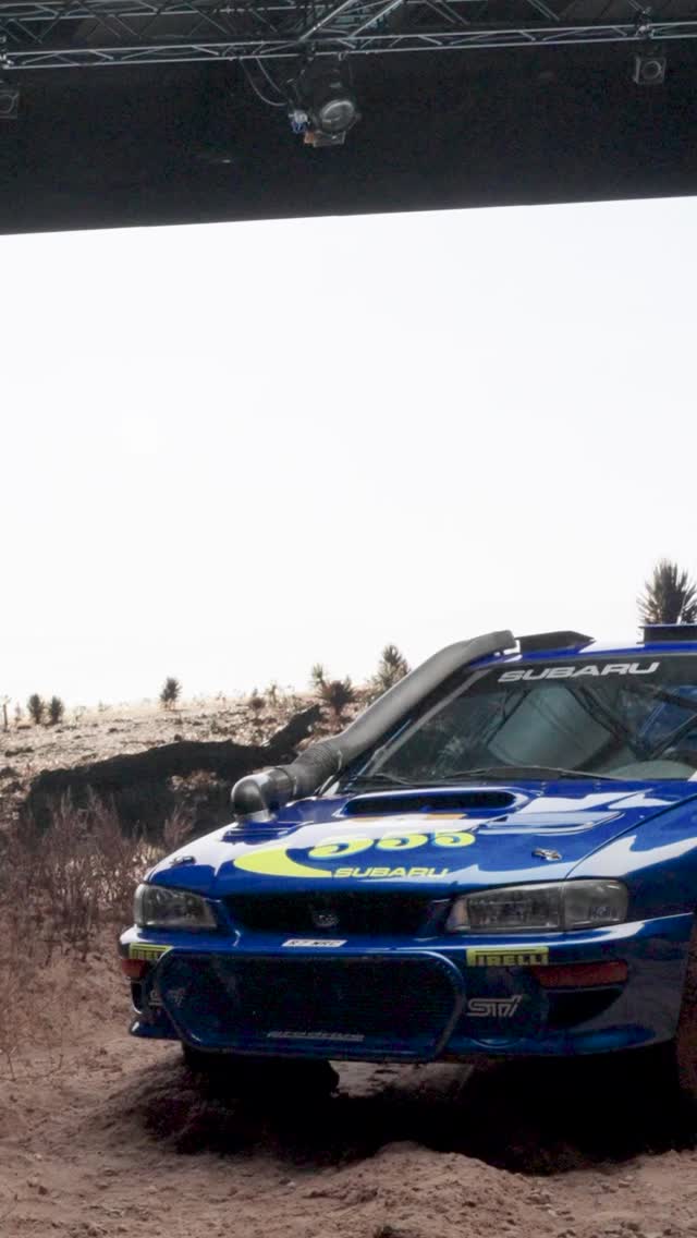 A blue Subaru rally car with yellow decals, including the number 555, is parked on a dirt surface under a structure, with a snowy and grassy landscape in the background.