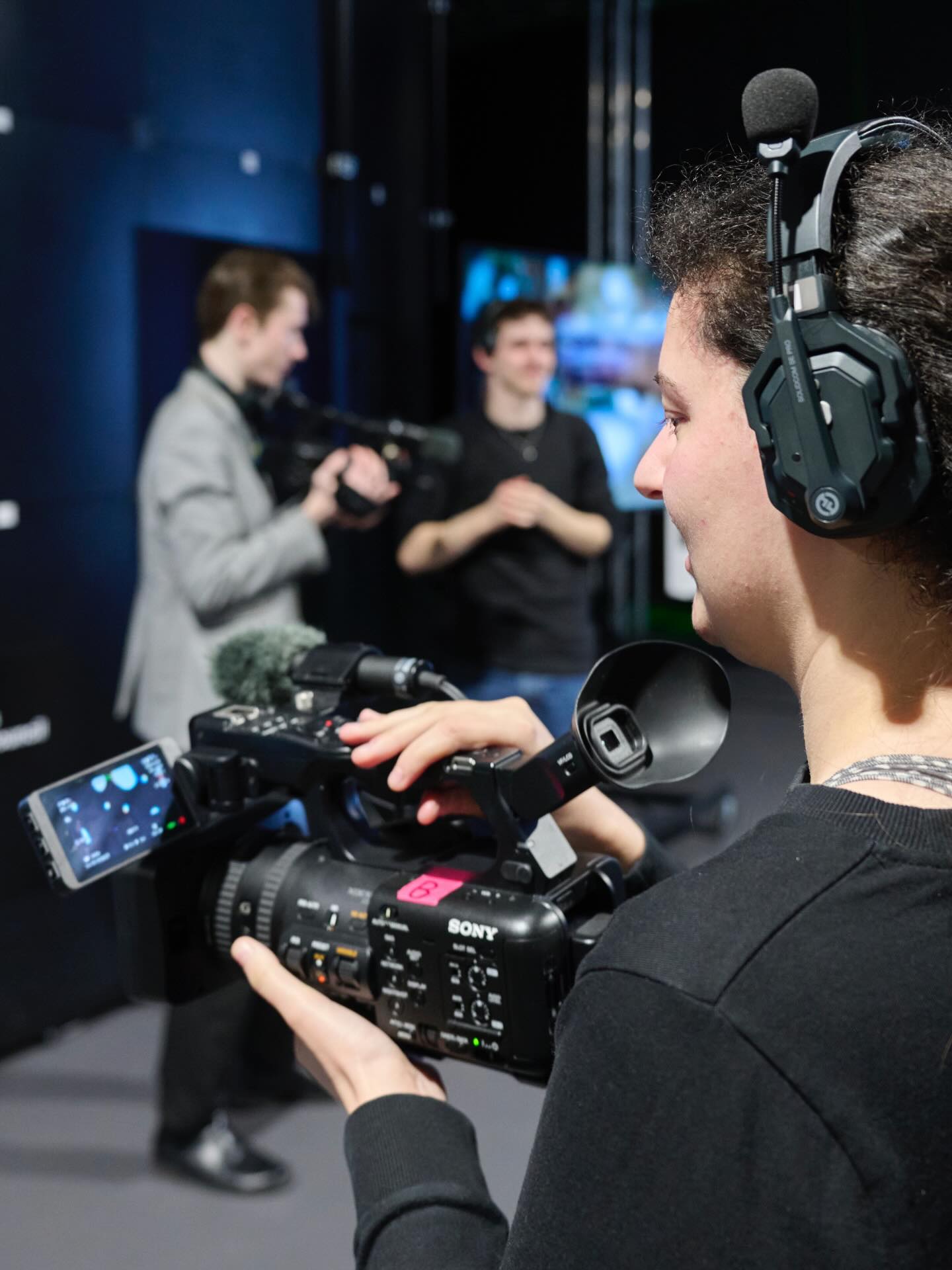 A person wearing a headset operates a professional video camera, focusing on two people in the background who are also filming in a studio environment.