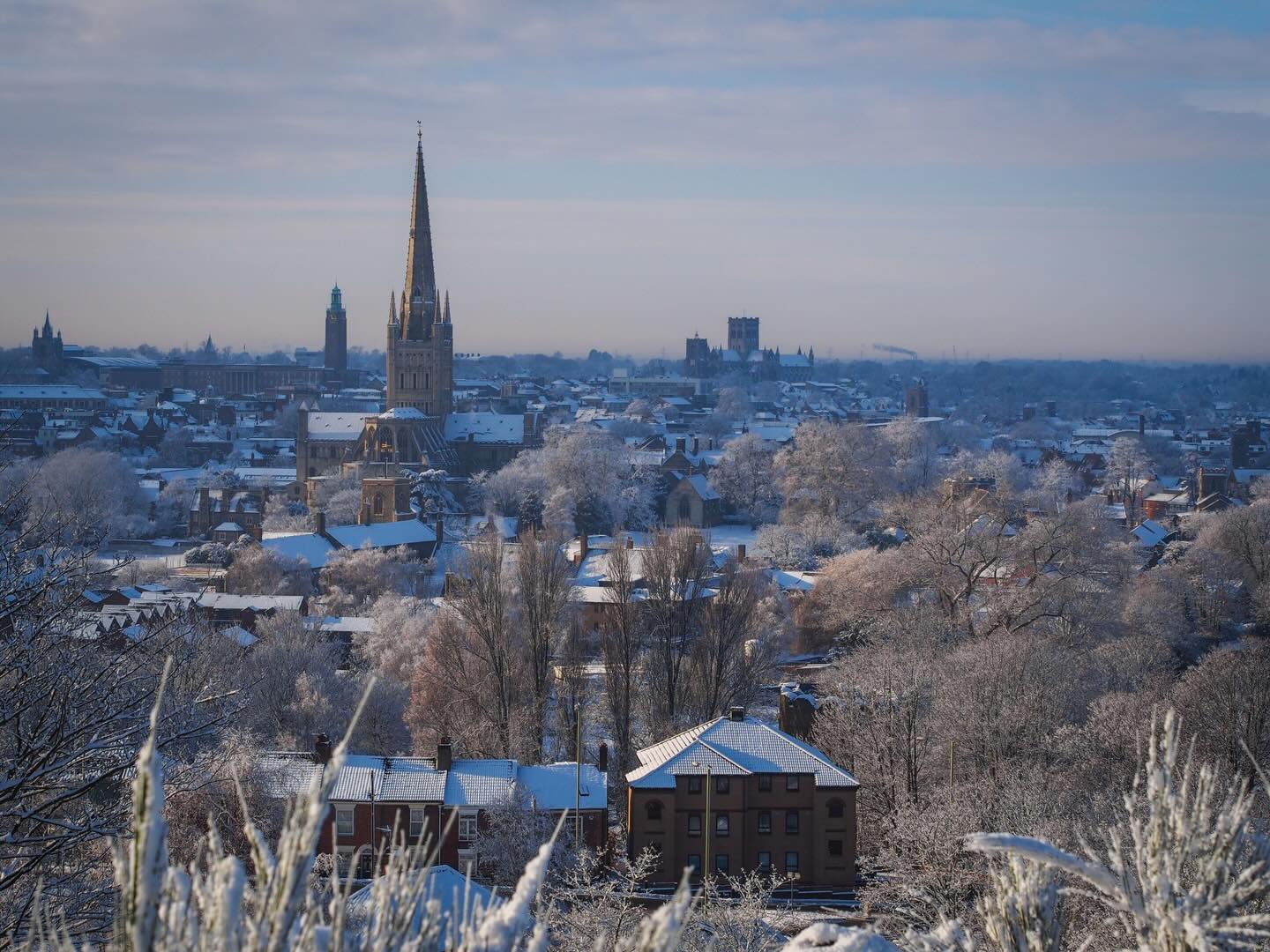 A winter cityscape shows rooftops, trees, and church spires blanketed in snow under a pale blue sky. The scene is peaceful and bright, with frosted vegetation in the foreground.