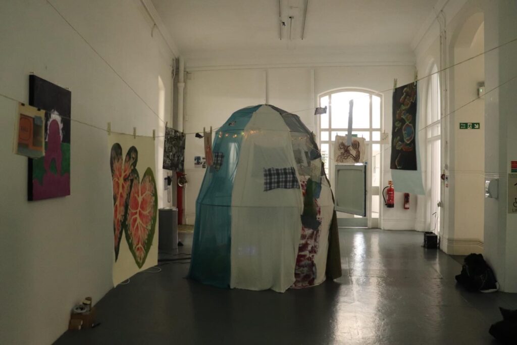 A fabric tent-like installation stands in a white-walled gallery, surrounded by colorful paintings and artwork hanging on string and walls. Large windows at the back let in natural light, brightening the exhibition space.