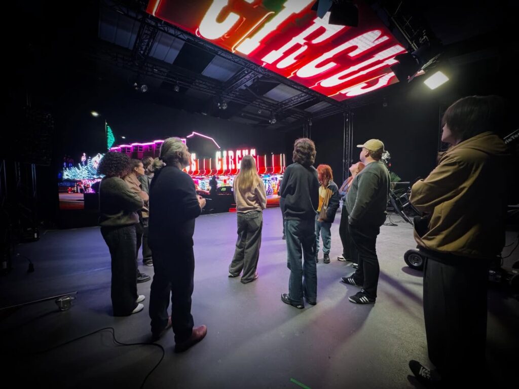 A group of people stand indoors facing a brightly lit stage with a large "CIRCUS" sign overhead and colorful lights in the background, suggesting a circus or performance setting.
