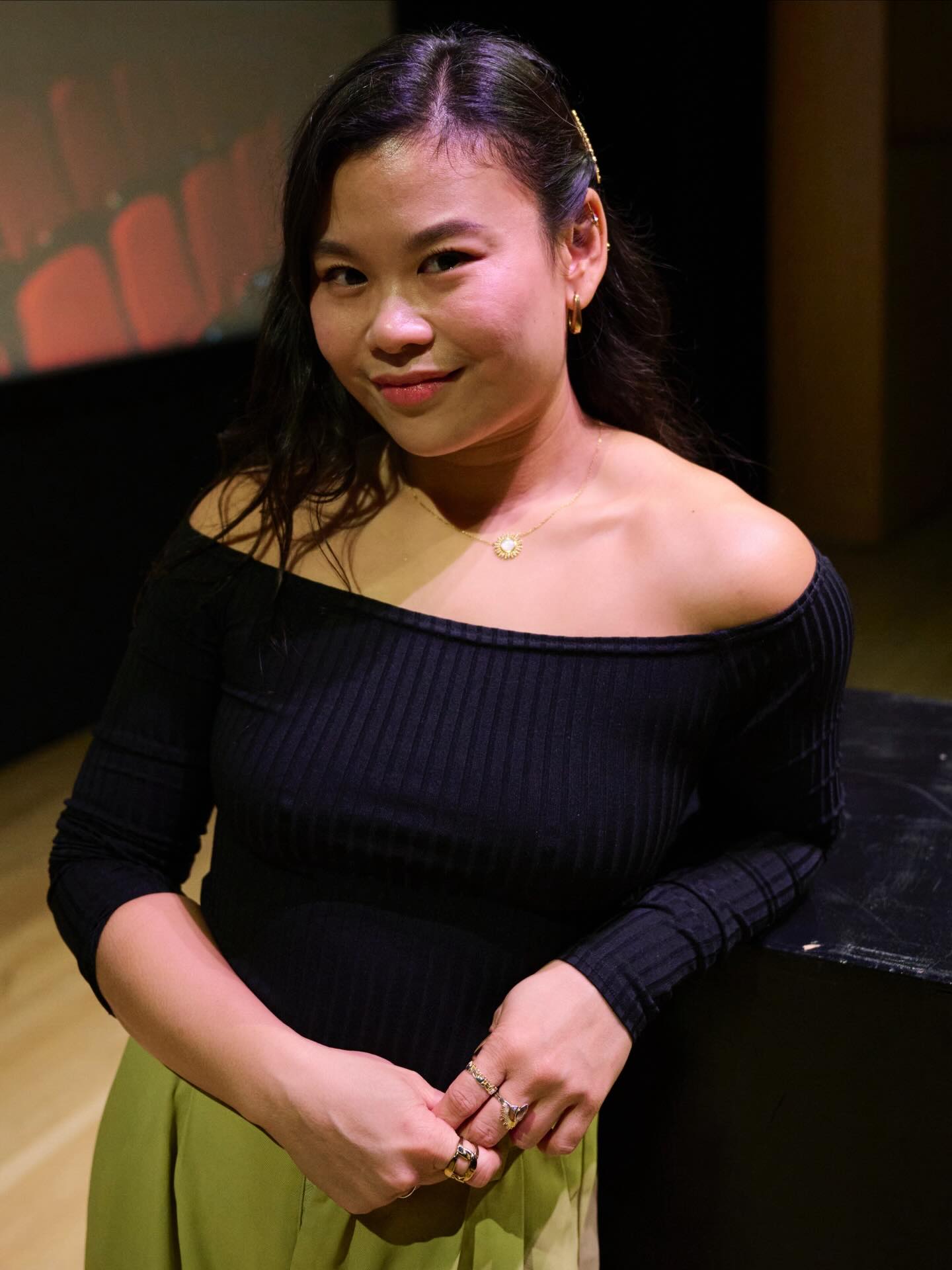 A woman with long dark hair, wearing a black off-the-shoulder top and light green pants, stands indoors leaning on a dark surface. She smiles slightly at the camera, with a blurred theater background behind her.