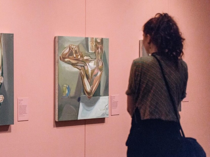 A person with curly hair and a shoulder bag stands in front of a surreal-style painting in an art gallery with pink walls, closely observing the artwork.