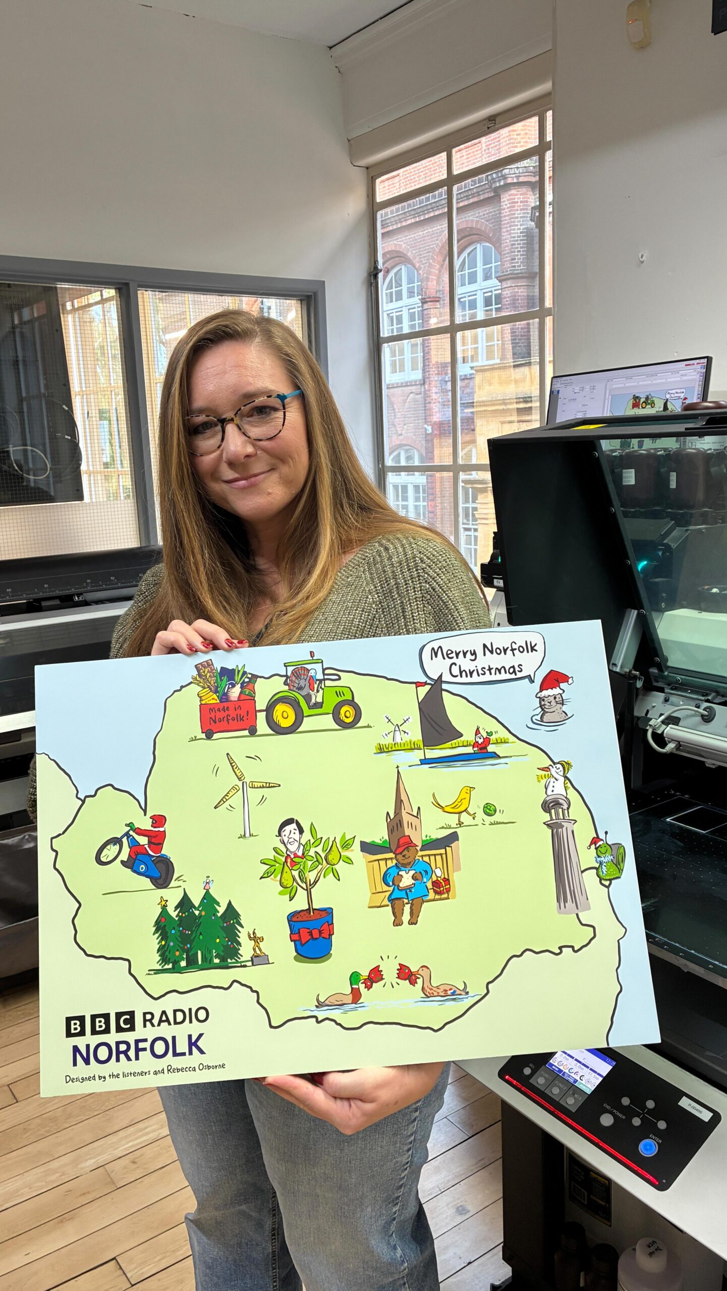 A woman with long hair and glasses smiles while holding a colorful illustrated map labeled "Merry Norfolk Christmas" and "BBC Radio Norfolk," featuring festive scenes and local landmarks.