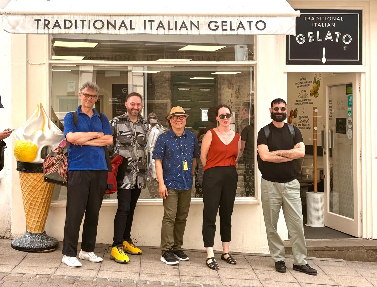 Five people stand smiling in front of a shop with a sign reading "Traditional Italian Gelato." A large decorative ice cream cone is by the door. The group appears relaxed and happy on a sunny day.