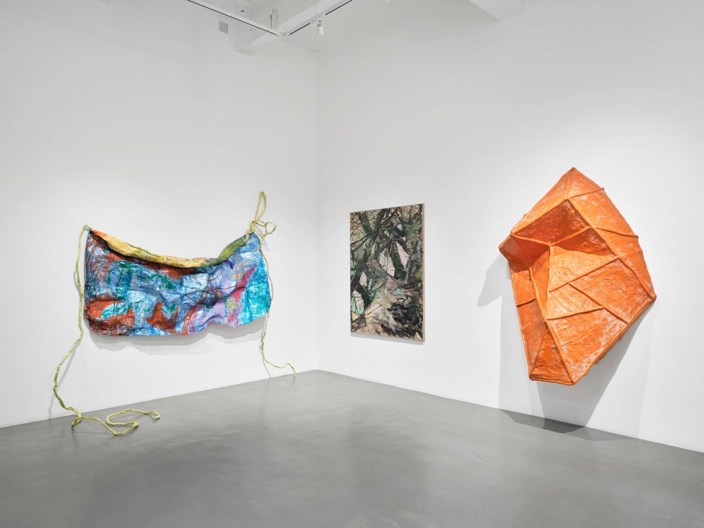 A white-walled gallery displays three artworks: a colorful, draped textile with yellow rope on the left, a textured painting in the center, and a large, angular orange sculpture mounted on the right wall.
