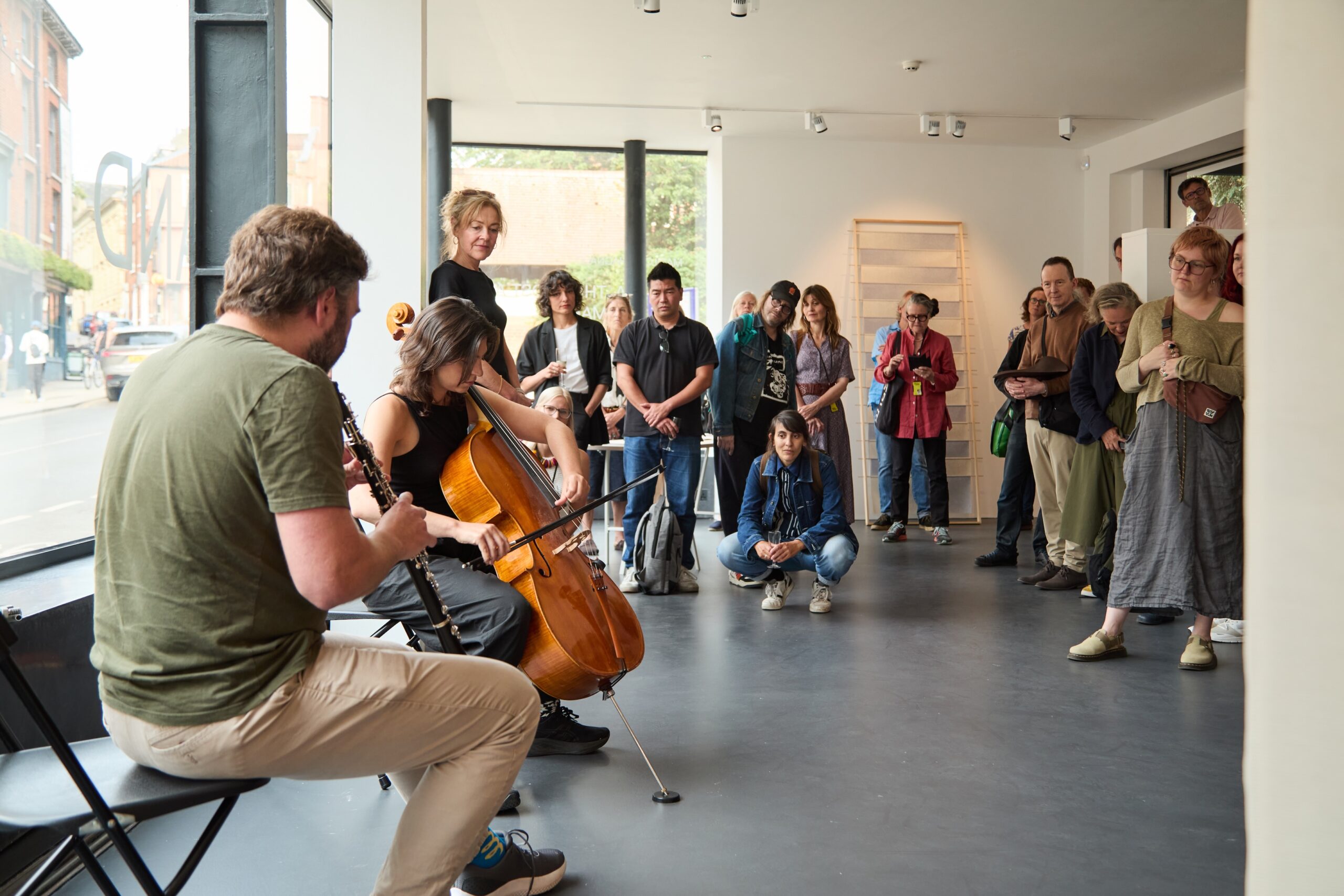 A group of people stand and sit in a modern gallery space, watching two musicians perform with a cello and clarinet near large windows, while some audience members take photos or listen attentively.