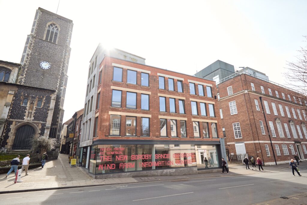 A modern brick building with large windows sits on a street corner next to an old church with a clock tower. People walk on the sidewalk, and a red digital sign is visible on the building’s ground floor.