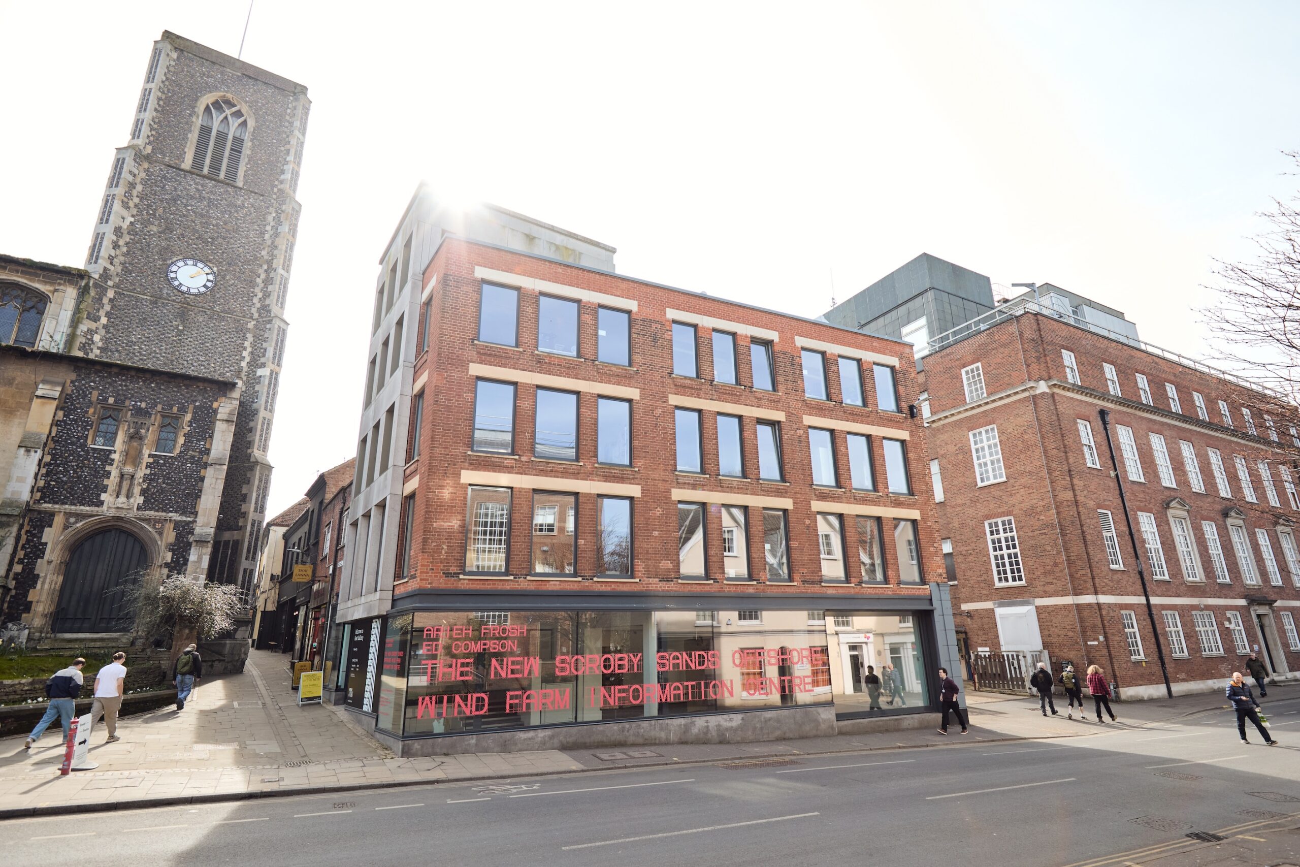 A modern brick building with large windows sits on a street corner next to an old church with a clock tower. People walk on the sidewalk, and a red digital sign is visible on the building’s ground floor.