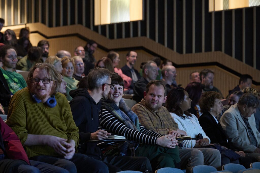 A diverse group of people sit in tiered seating at an indoor event, chatting and waiting for it to begin. Some are smiling and talking, while others look forward or are engaged with their phones.
