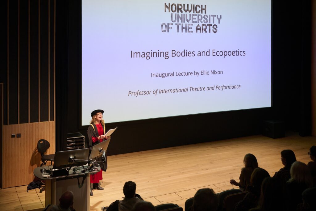 A person in academic dress stands at a podium delivering a lecture in a modern auditorium. A presentation slide behind them reads “Norwich University of the Arts: Imagining Bodies and Ecopoetics, Inaugural Lecture by Elin Nixon.”.