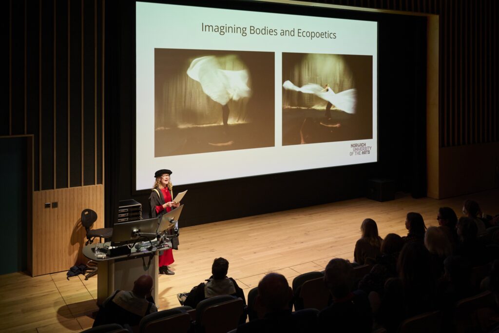 A person in academic attire stands at a podium presenting to an audience. On the screen behind them are two blurred images of a dancer and the title “Imagining Bodies and Ecopoetics.”.