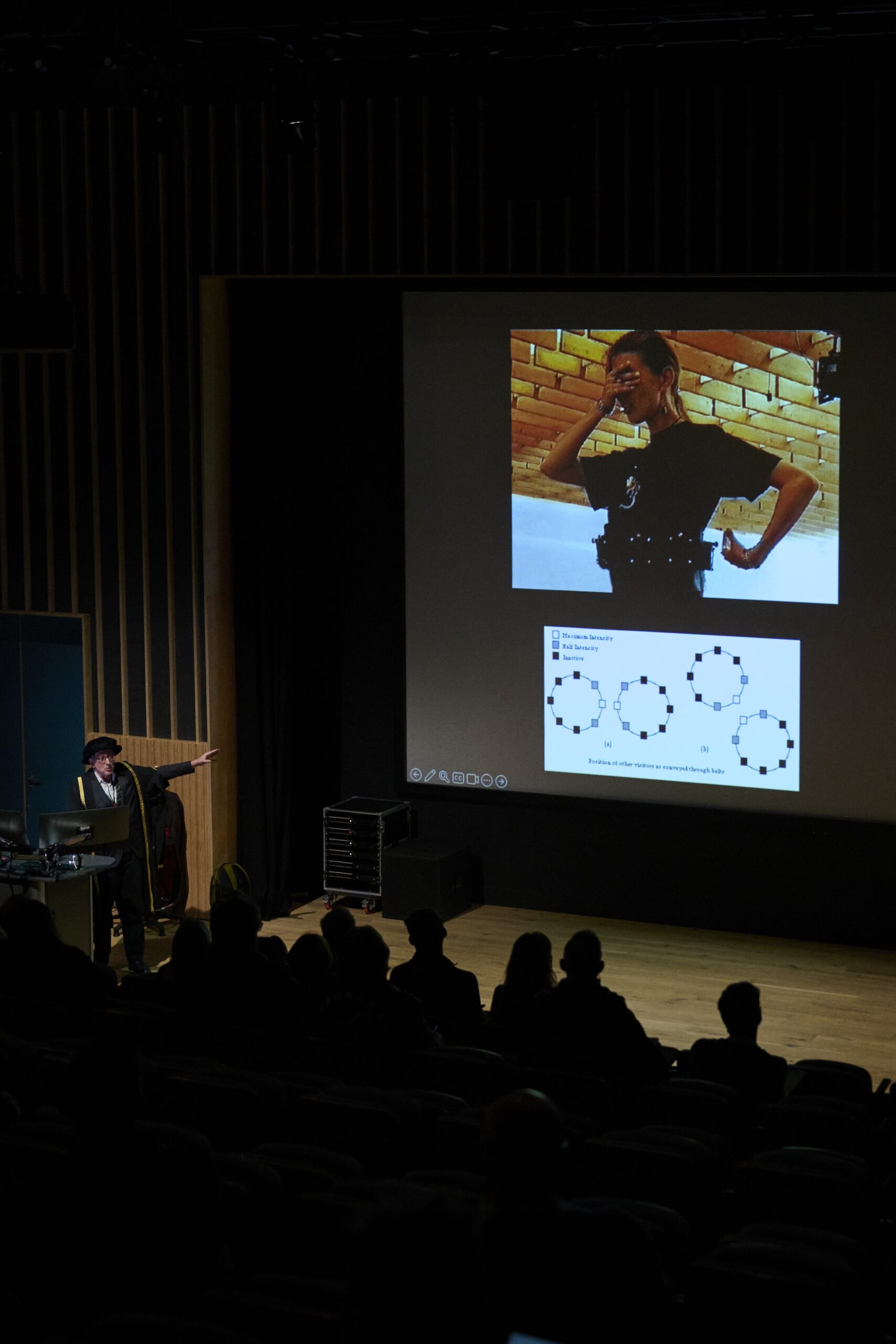 A person gives a presentation in a dark auditorium, pointing at a projected screen showing a photo of someone posing and diagrams of circles. Audience members are silhouetted in the foreground.