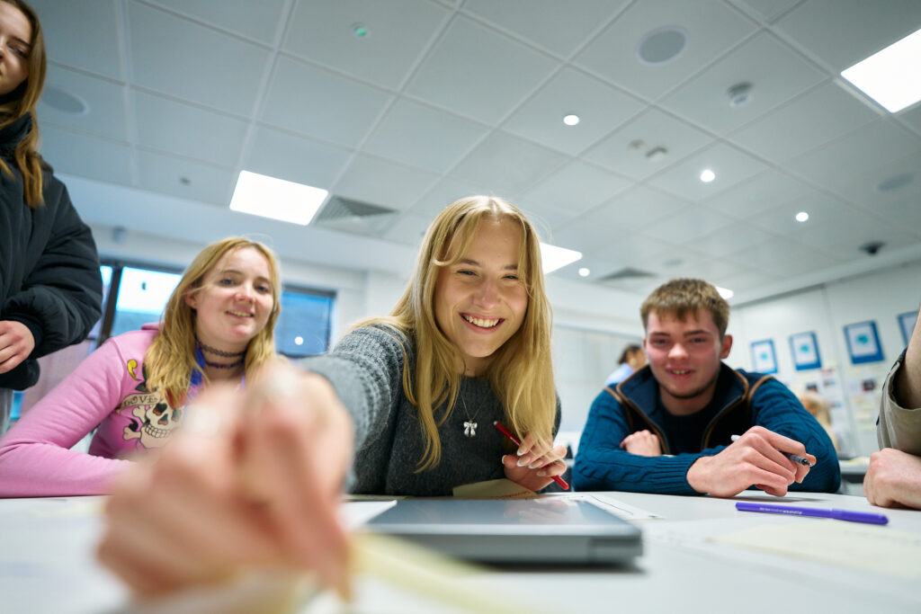 A group of smiling young people sit at a table in a bright classroom, engaging in an activity together. One person in the foreground reaches forward with a pen, appearing cheerful and involved.