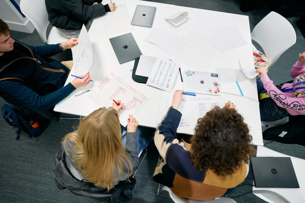 Five students sit around a white table, collaborating and writing notes on paper and whiteboards. Laptops, documents, and notebooks are scattered across the table. The image is taken from above.