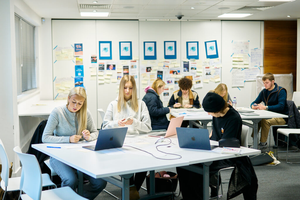 A group of students sits at tables in a classroom, working on laptops and writing on paper. The walls are covered with notes, drawings, and photos. The atmosphere is focused and collaborative.