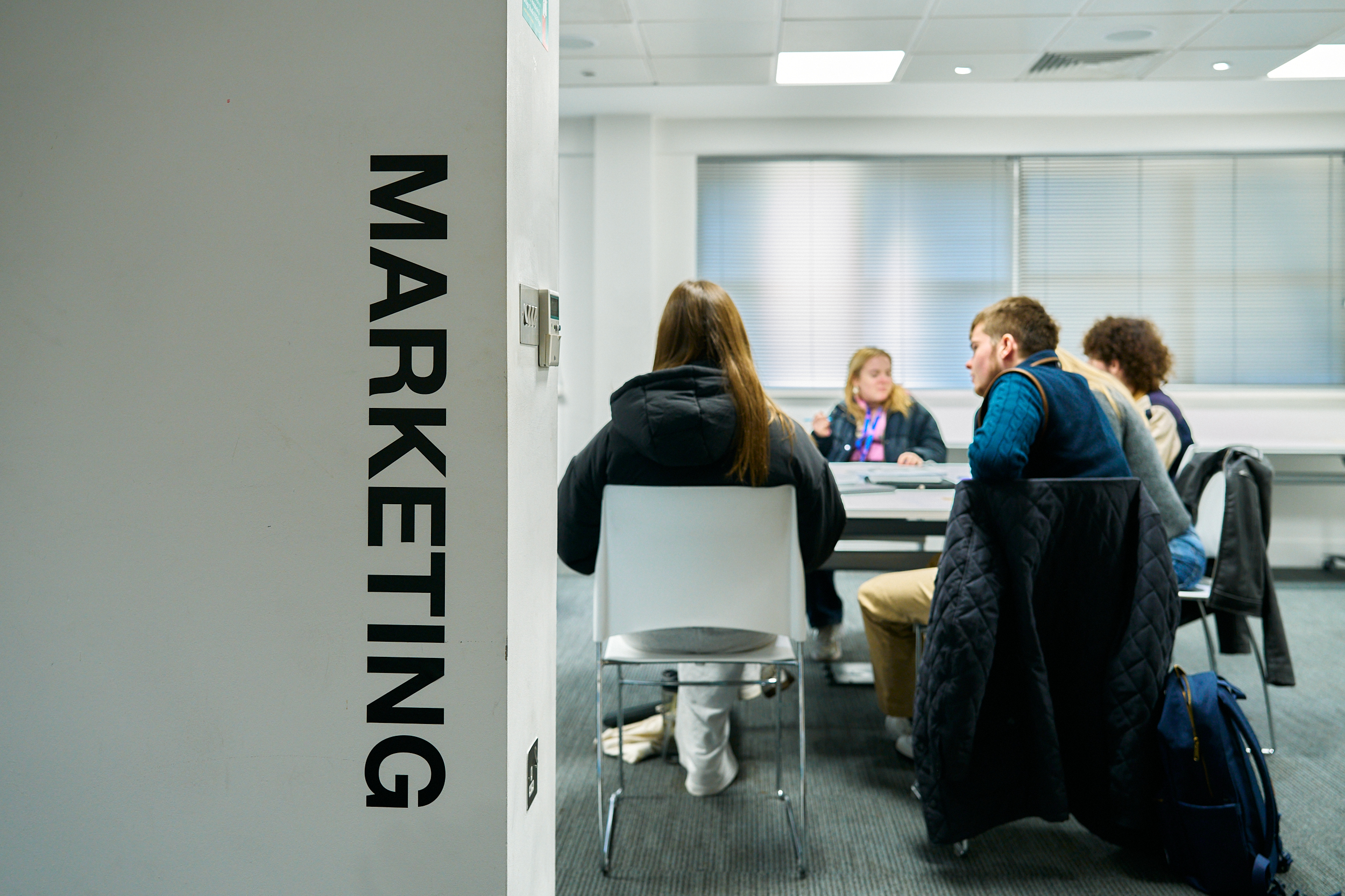 Four people sit around a table in a modern university room. The word "MARKETING" is prominently displayed on a wall in the foreground.