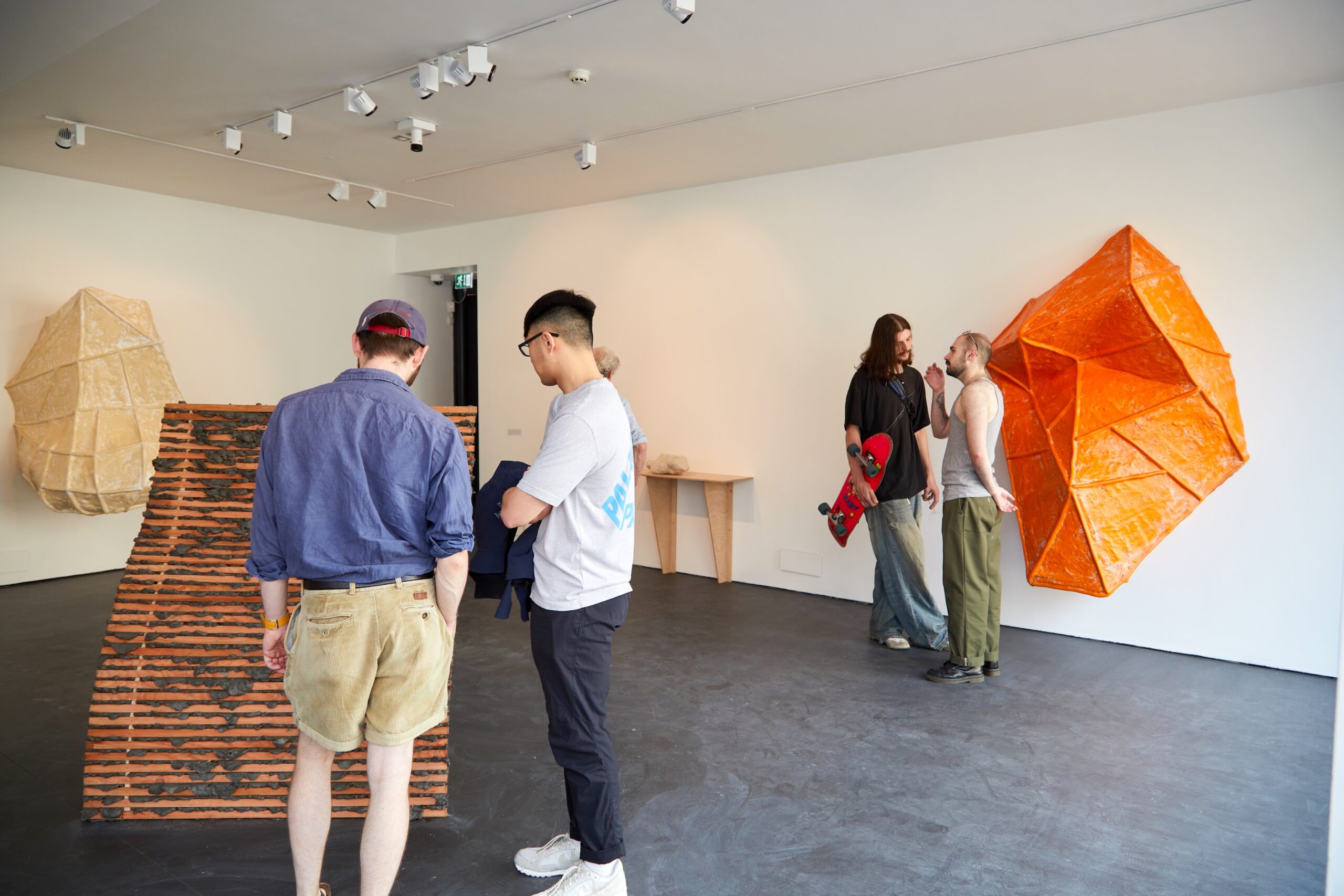 People view abstract sculptures and artwork in a modern gallery with white walls and dark floors. Two men observe art closely, while a small group converses near bright orange geometric wall art.