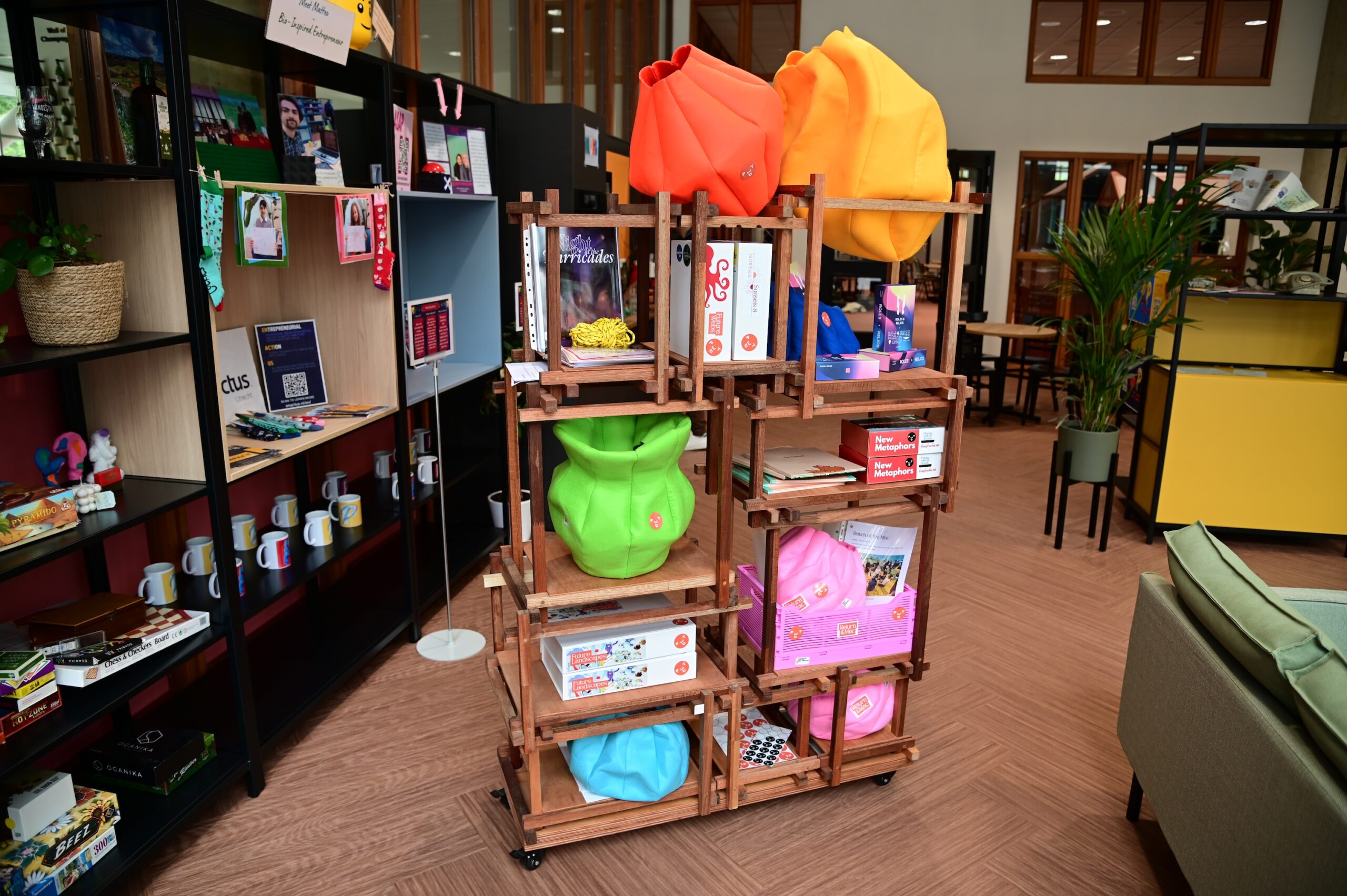 A wooden display shelf holds colorful plush hoodies, board games, mugs, and various gifts in a modern, brightly-lit store with other shelves and plants in the background.