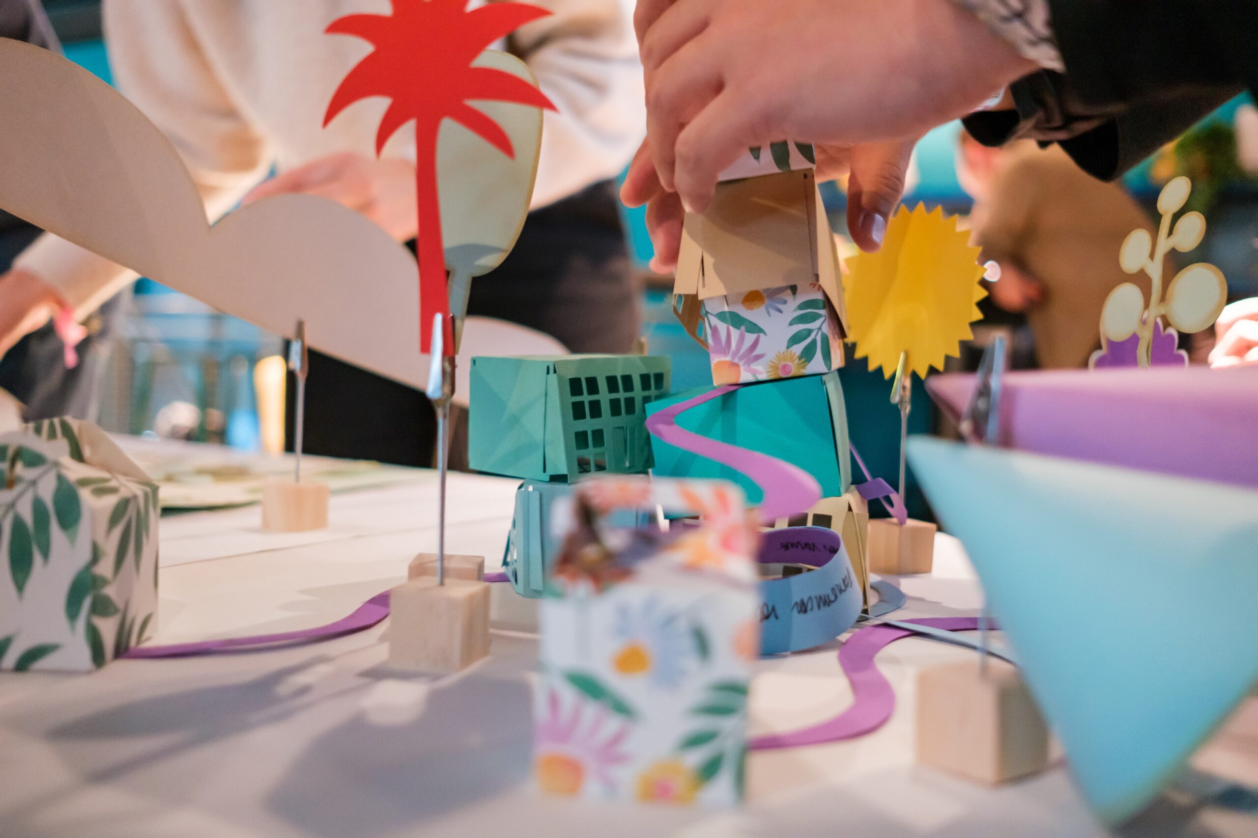 Several hands arrange colorful, patterned paper shapes and cardboard cutouts on a table, creating a playful, artistic scene with trees, buildings, and nature-themed decorations.