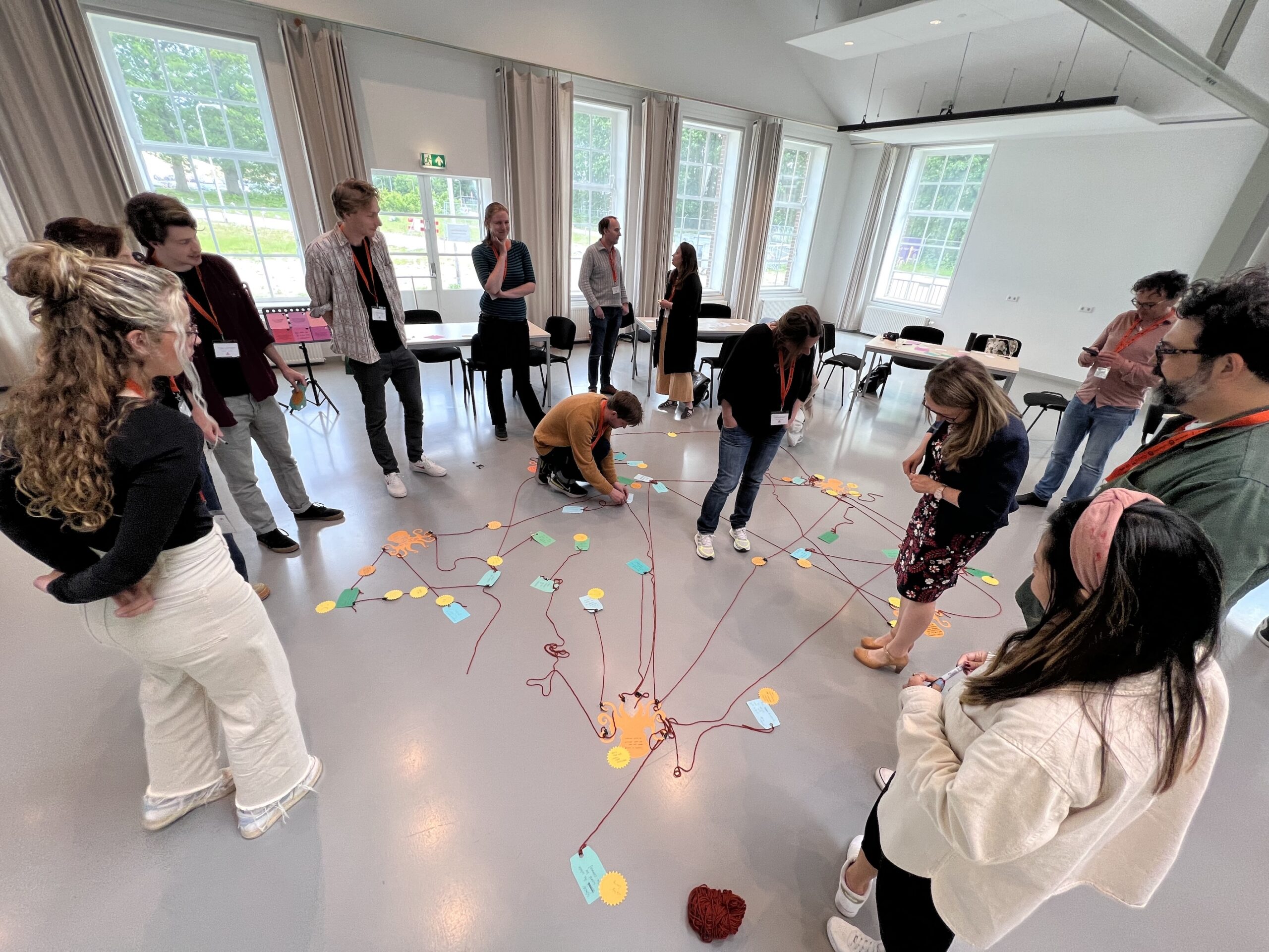A group of people stands in a circle in a bright room, connecting colored cards on the floor with red yarn. Some are bending down to tie yarn, while others watch or hold string, engaged in a collaborative activity.