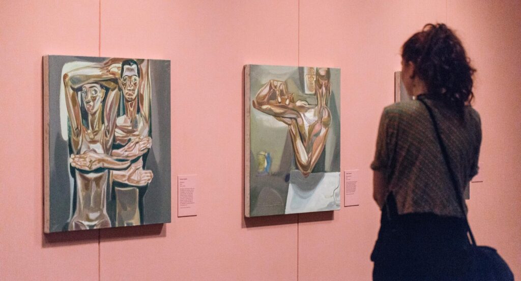 A person with curly hair stands in front of two abstract paintings of human figures displayed on a pink wall in an art gallery.