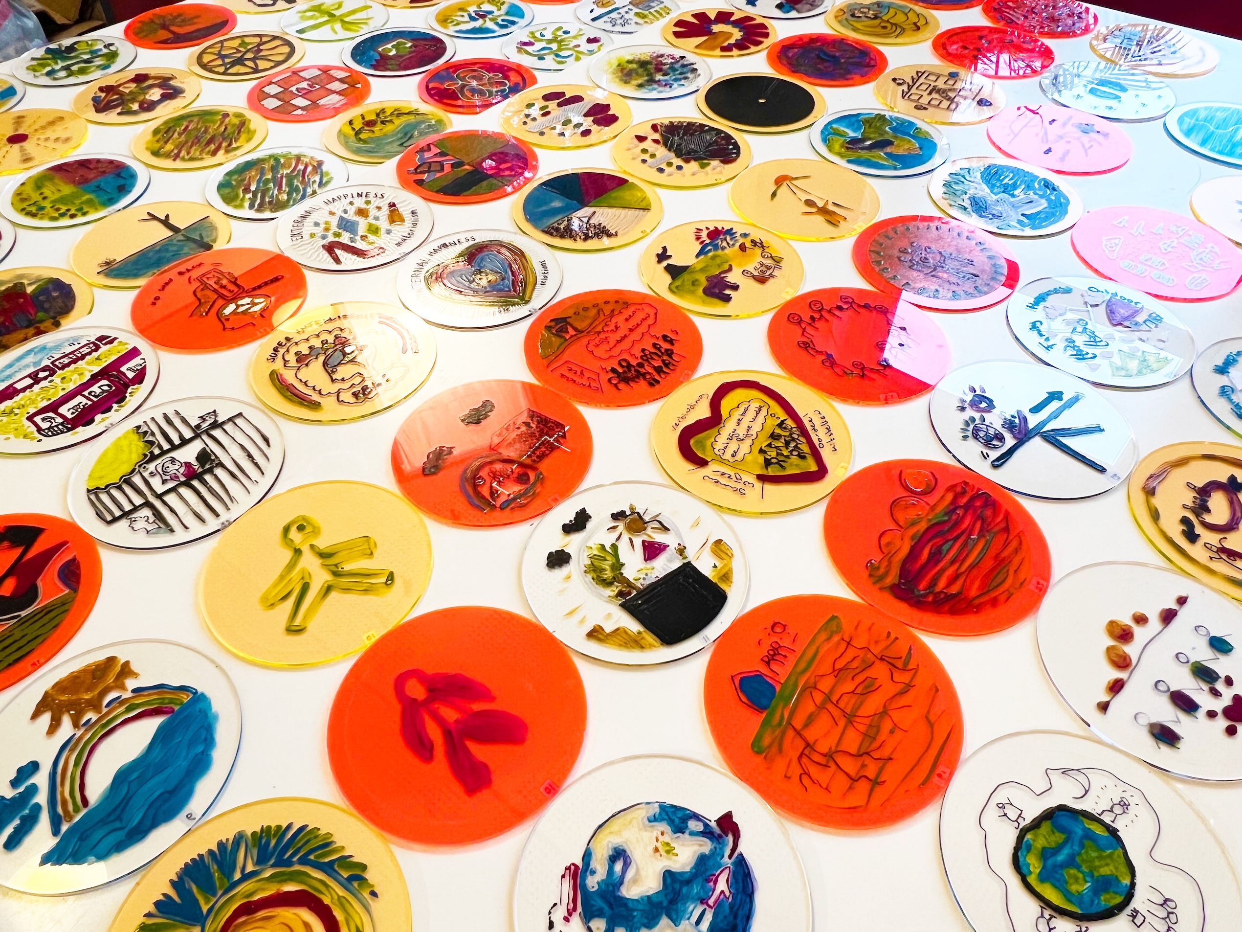 Colorful, circular art pieces with various drawings and designs are spread across a white surface, featuring different subjects like people, animals, hearts, and abstract patterns.
