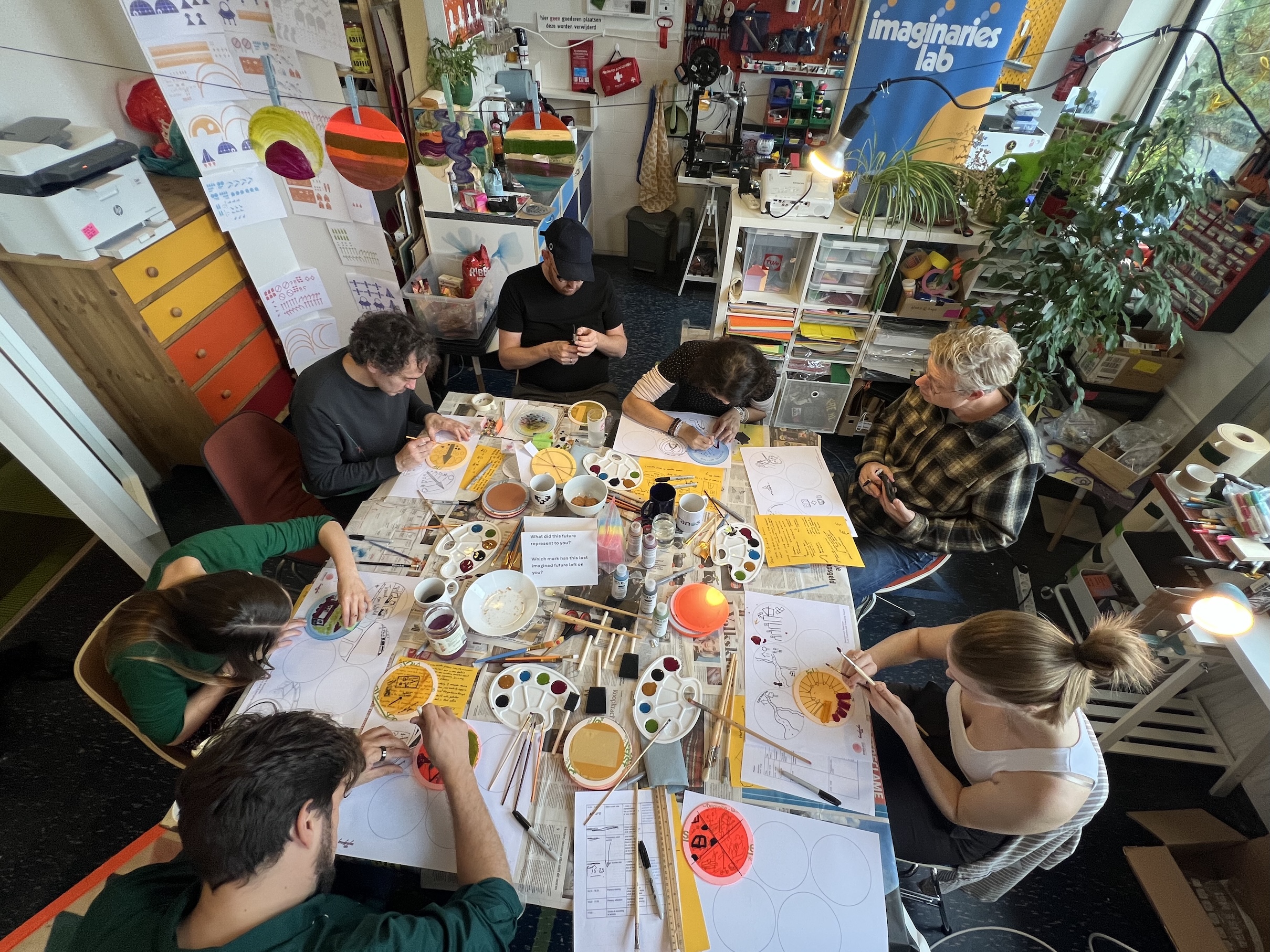 A group of people sit around a table filled with art supplies, papers, and colorful paints, working on creative projects in a bright, busy studio space decorated with art and signs.