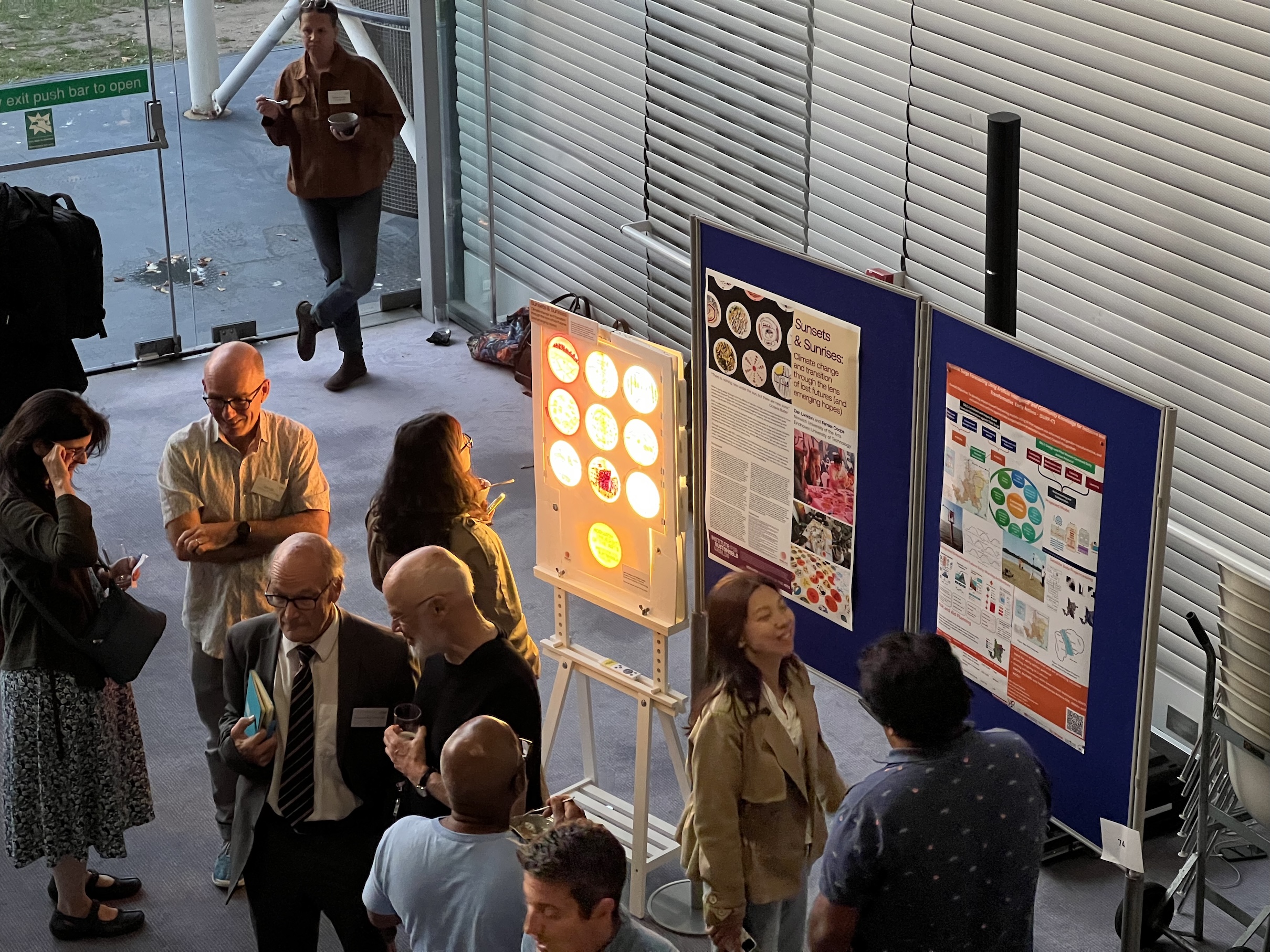 Overhead view of people conversing at an indoor event near posters and a brightly lit display board, with windows and an exit door visible in the background.