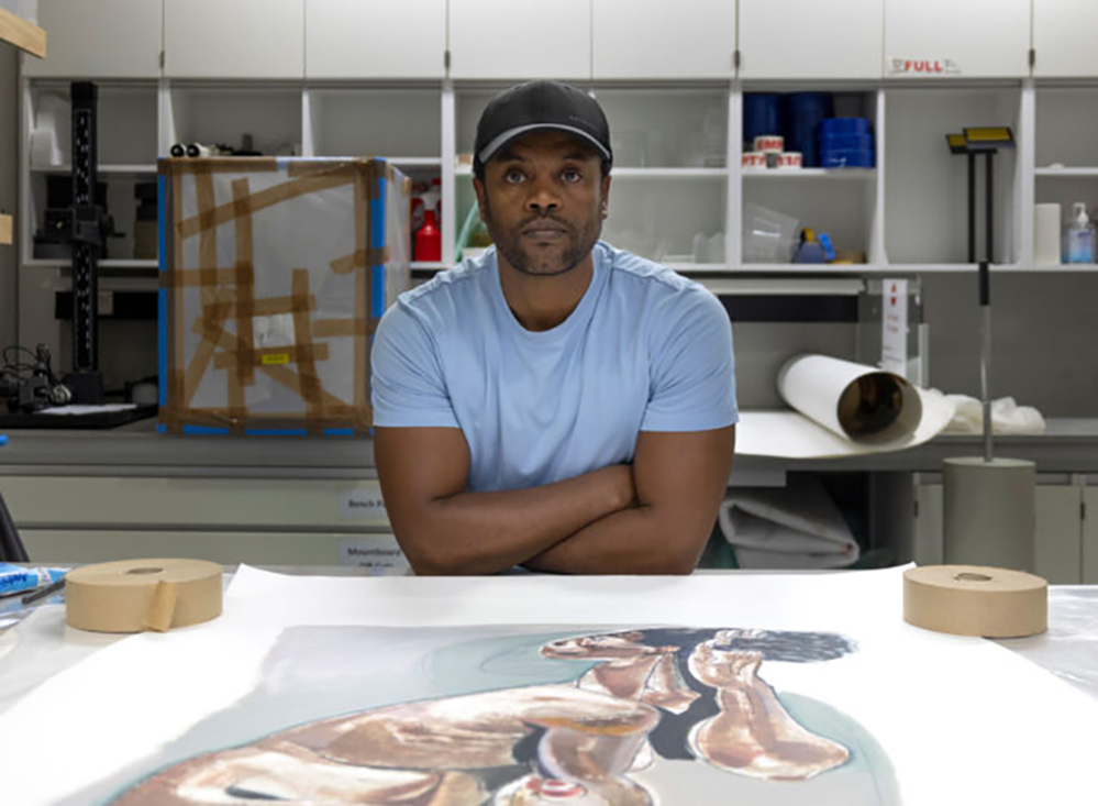 A man in a light blue shirt and black cap stands with his arms crossed, leaning over a table with an artwork of a woman. He is in a workshop or studio with shelves and supplies in the background.