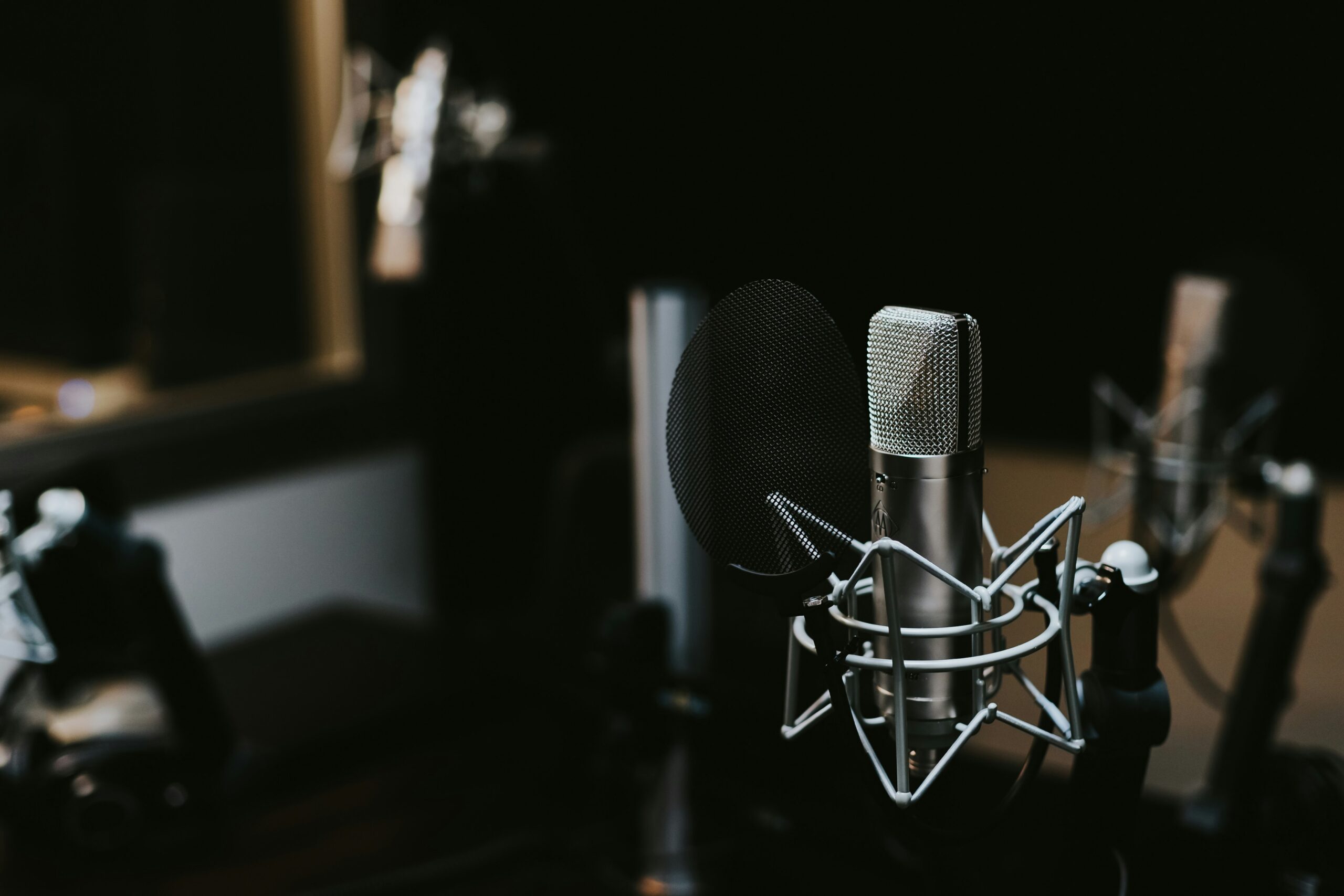 A close-up of a professional studio microphone with a pop filter, set up in a recording studio with other microphones blurred in the background. The lighting is dim, creating a focused atmosphere.