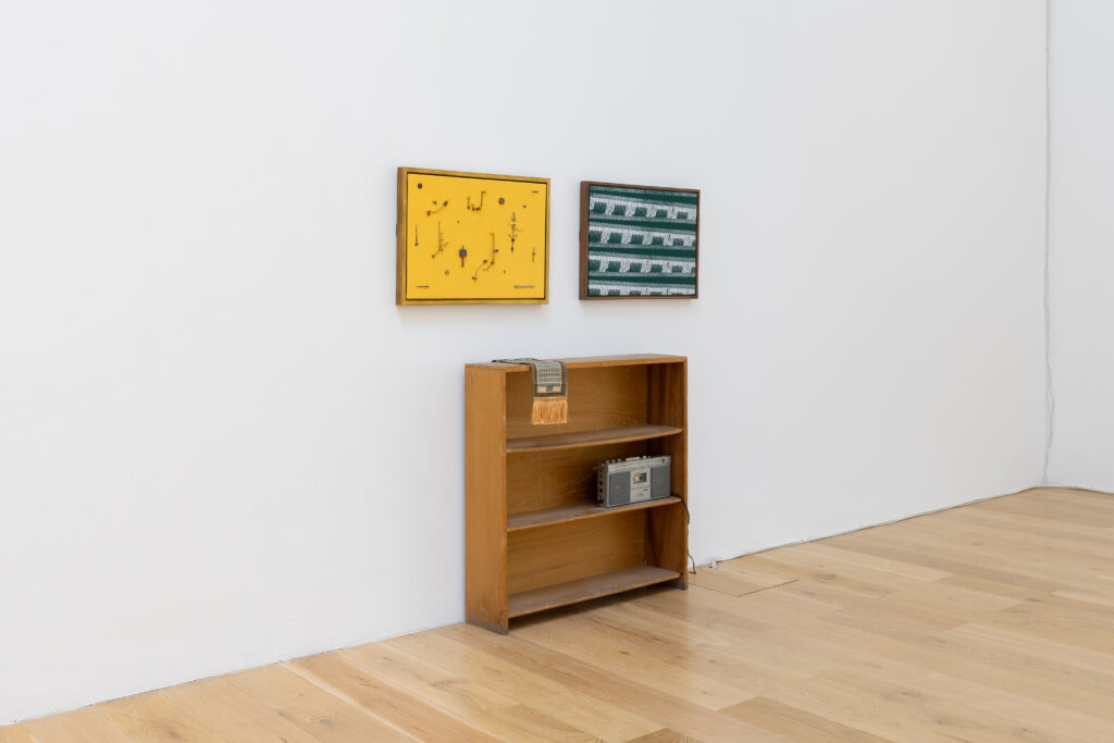 A small wooden bookshelf stands against a white wall on a wooden floor, with two framed artworks above it: one yellow with scattered black shapes, and one green with a geometric pattern. A metal box sits on a shelf.