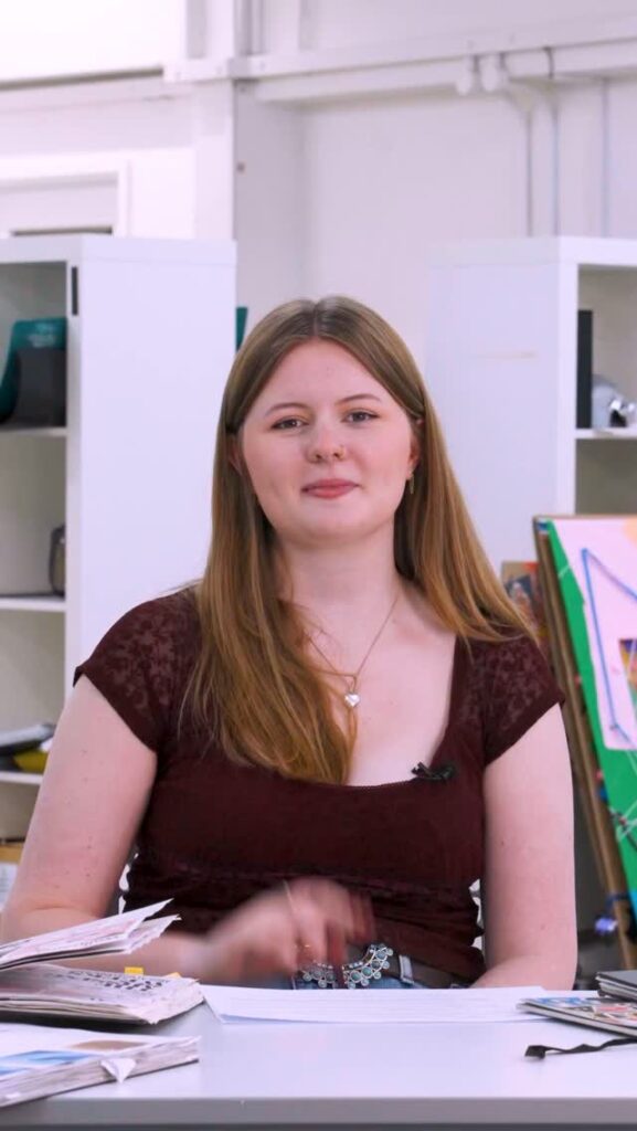 A young woman with long brown hair sits at a desk, smiling slightly. She is wearing a burgundy top and a necklace. Behind her are shelves, papers, and art supplies in a bright, modern room.