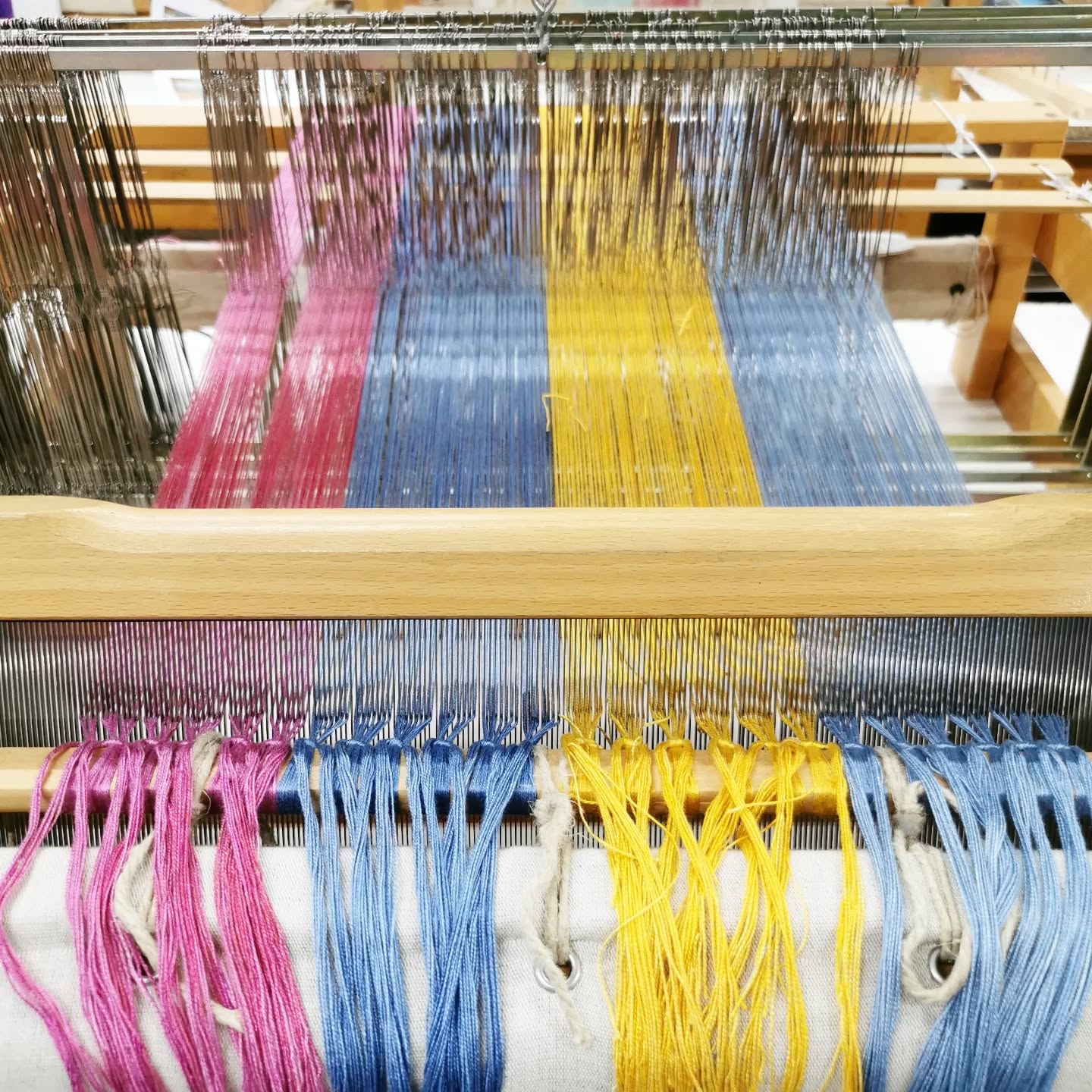 Close-up of a weaving loom with colorful threads in pink, yellow, and blue, arranged neatly and stretched through the loom’s heddles and reed, ready for weaving.