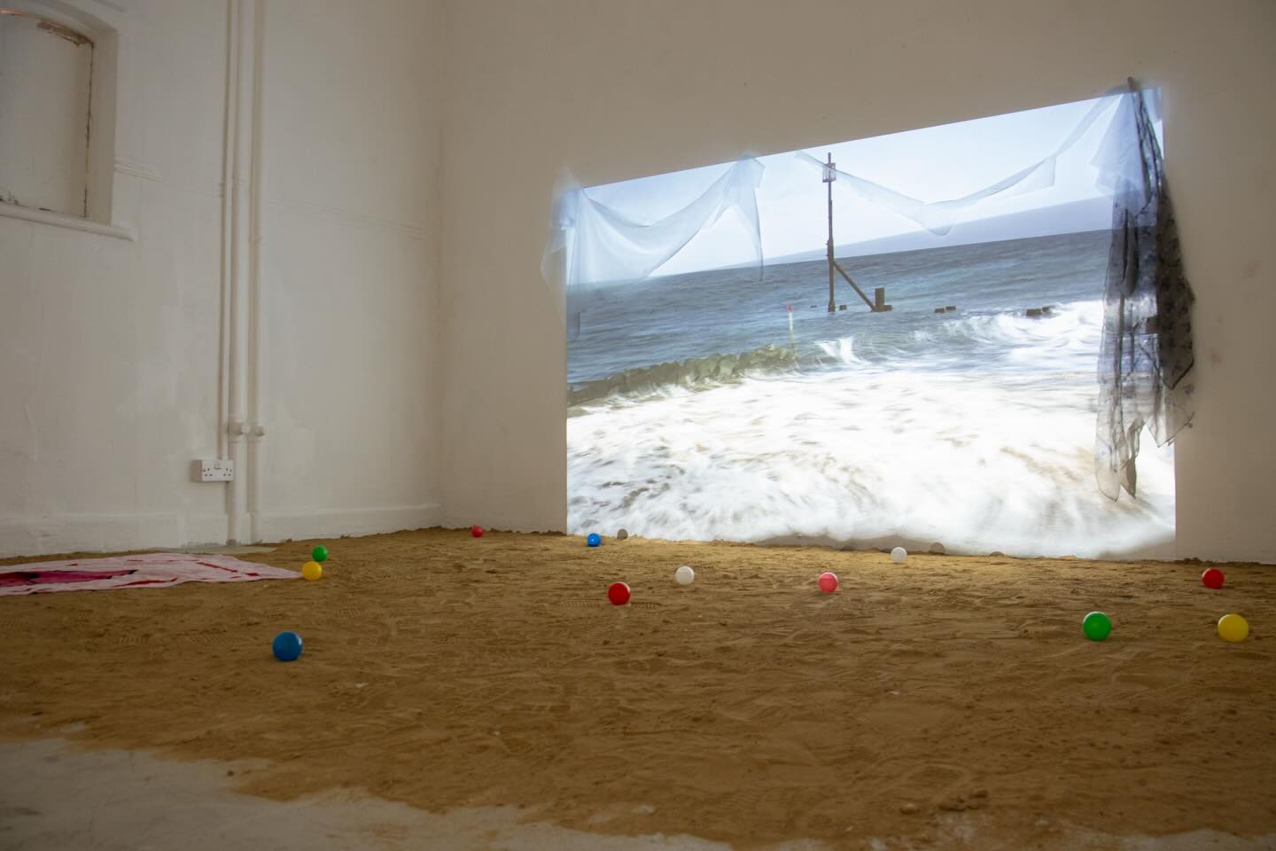 A room with a sandy floor, scattered colorful plastic balls, and a wall projection of an ocean scene with waves. Sheer fabric partially covers the projection, creating a beach-like atmosphere indoors.