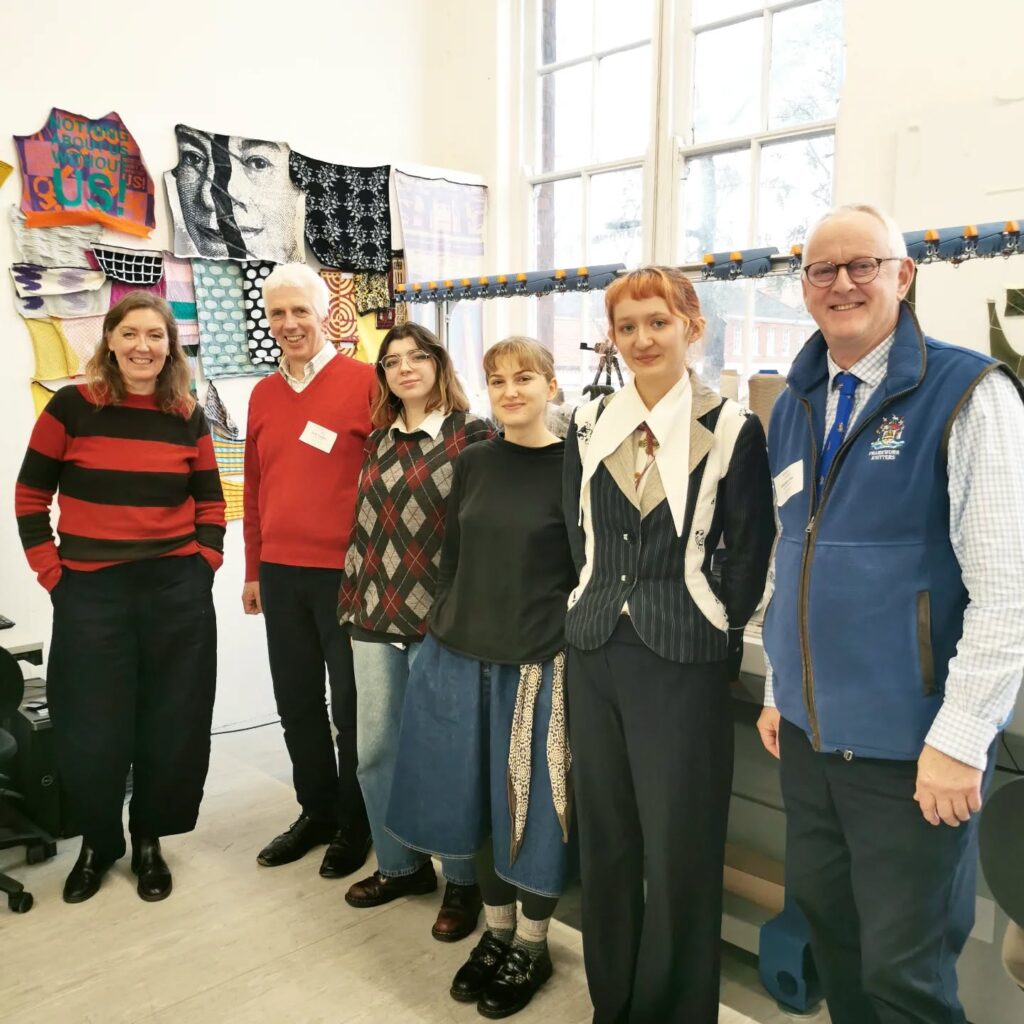 Six people stand in a row indoors, smiling at the camera. Colorful textile art and knitted pieces hang on the wall behind them, and large windows let in natural light.