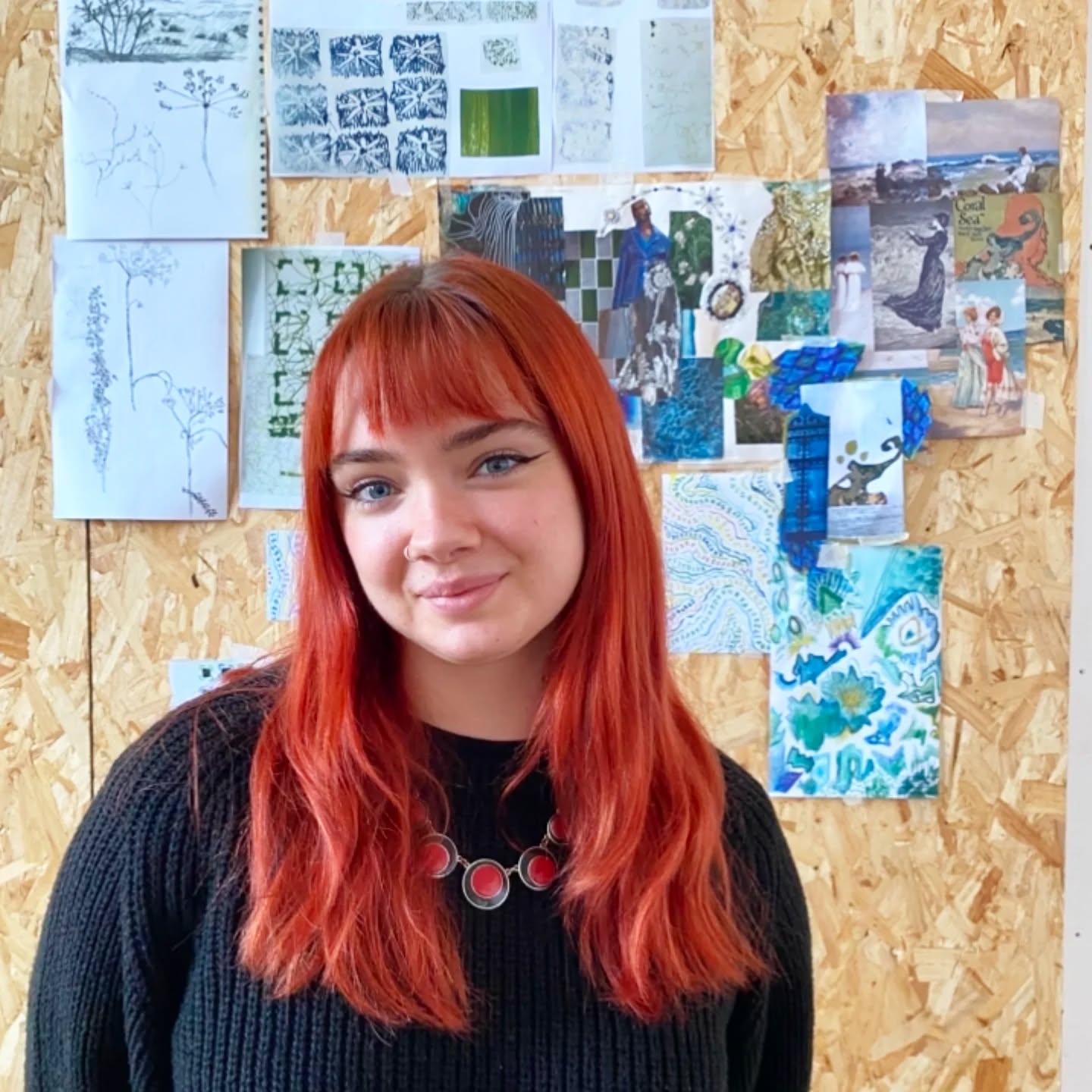 A woman with long red hair and bangs, wearing a black sweater and a red necklace, smiles in front of a corkboard wall covered with various art sketches and colorful pictures.