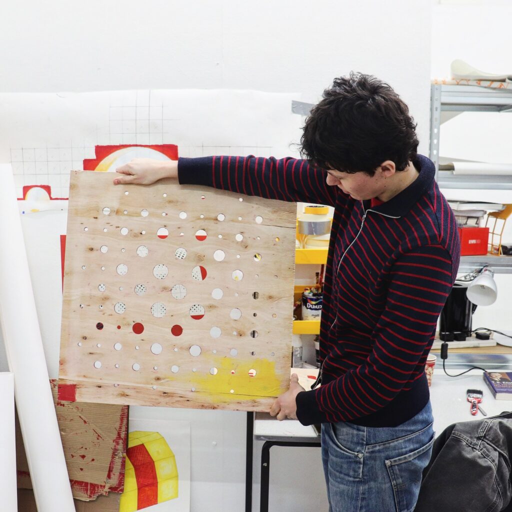 A person in a striped sweater holds up a wooden board with various circular holes cut out. They stand in a workshop, surrounded by art supplies, paper, and colorful objects.