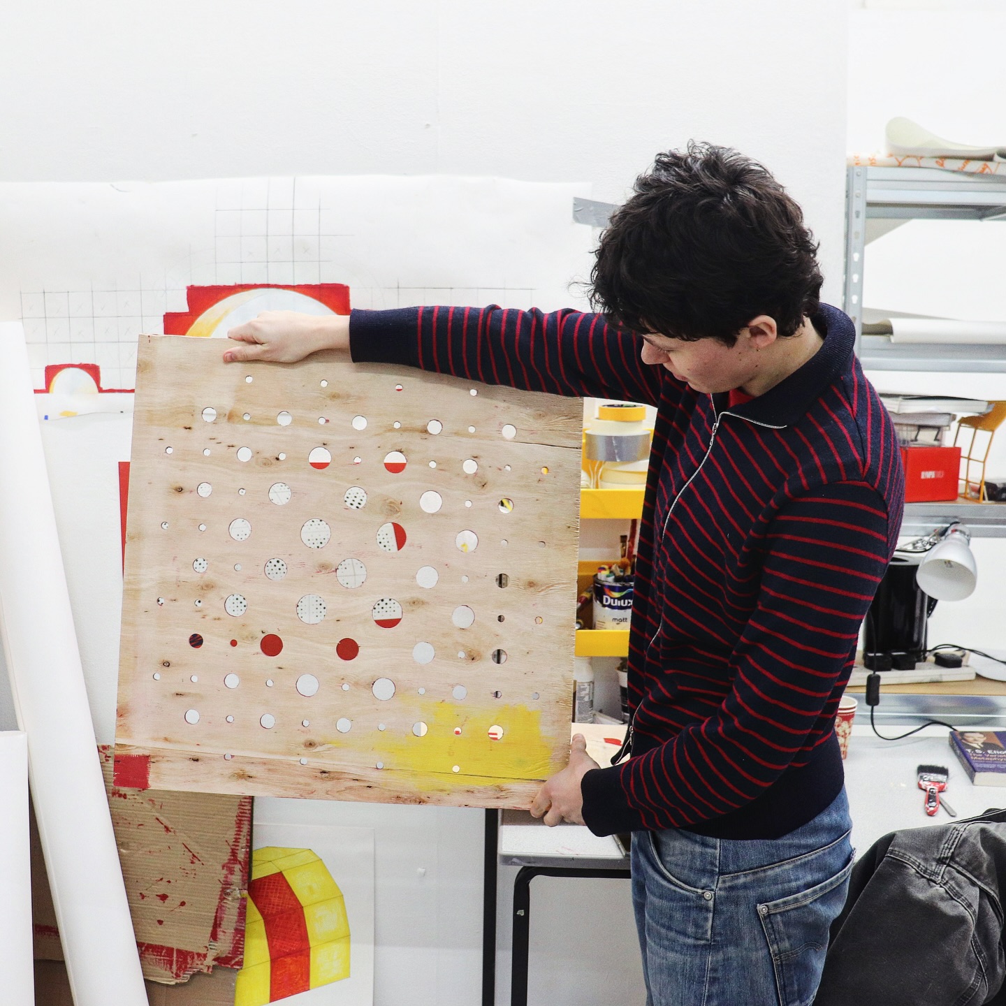 A person in a striped sweater holds up a wooden board with various circular holes cut out. They stand in a workshop, surrounded by art supplies, paper, and colorful objects.