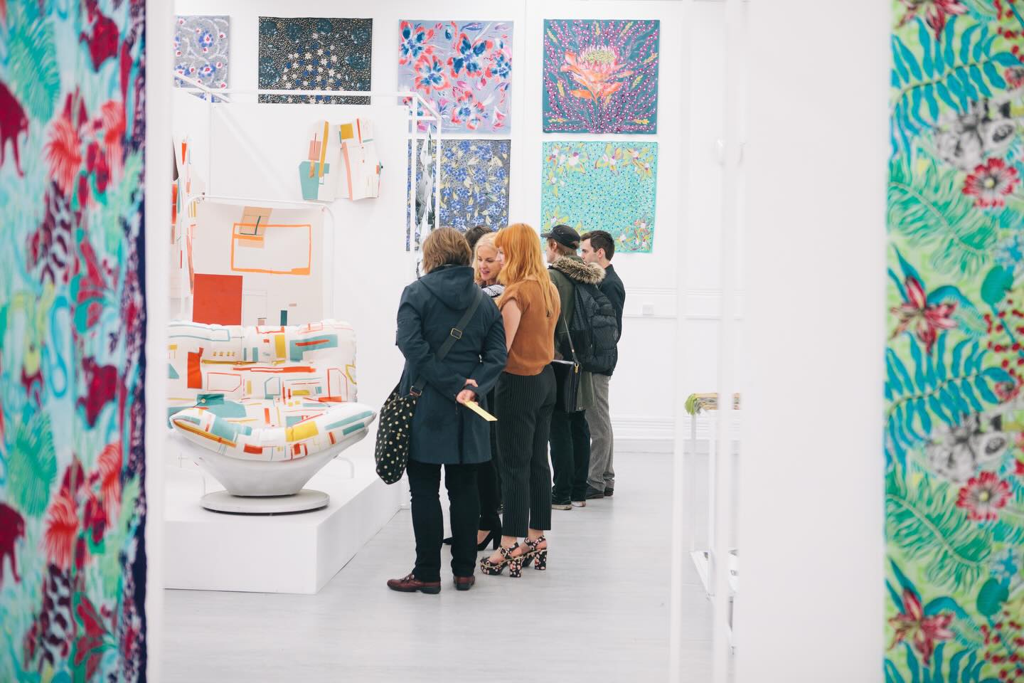 A group of people stand closely together, viewing colorful abstract sculptures in a modern art gallery. Patterned fabric panels and vibrant paintings decorate the bright, white-walled space.