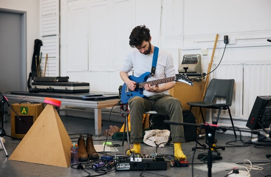 A man with dark hair plays an electric guitar while seated in a studio, surrounded by musical equipment, cables, and effects pedals. He wears a white shirt, yellow socks, and brown shoes sit nearby.