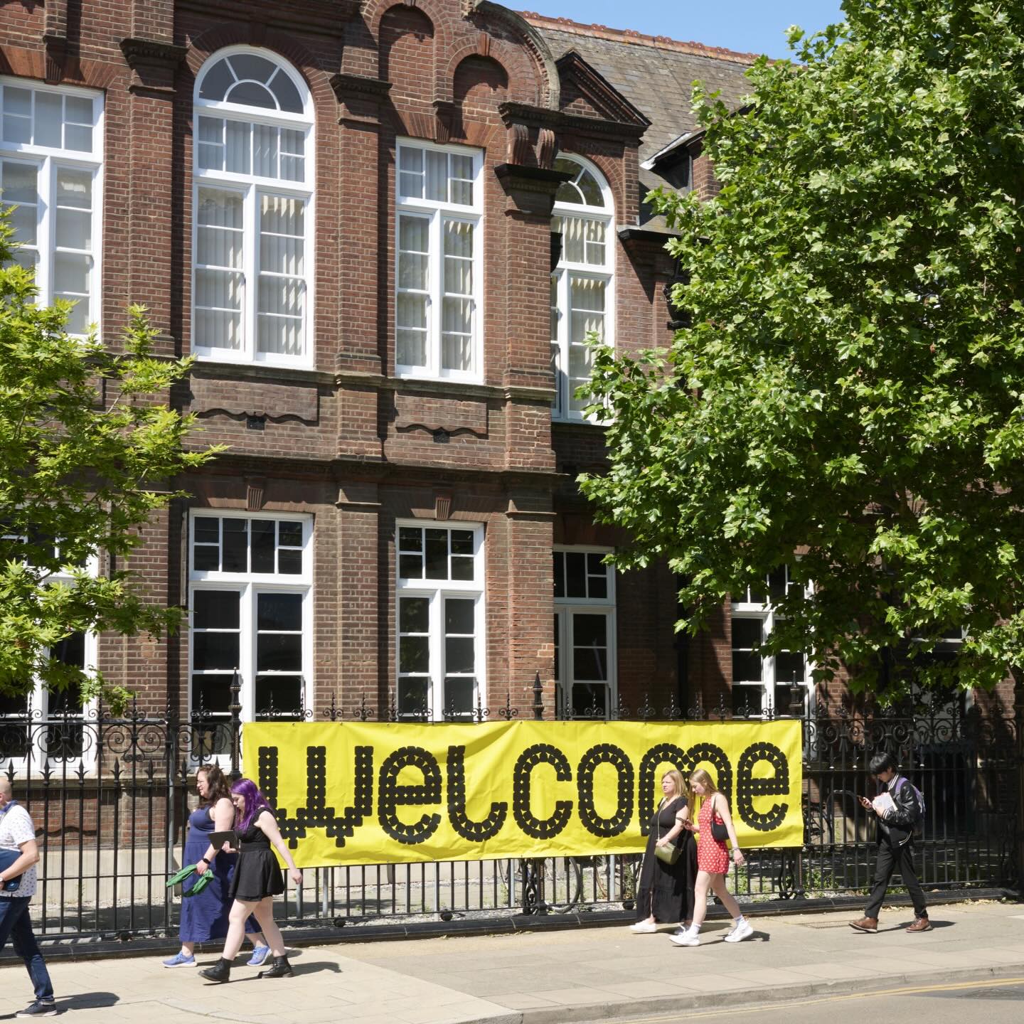 A large yellow “welcome” banner hangs on a black iron fence outside a brick building with tall windows. Several people walk past on the sidewalk, and green trees provide shade.