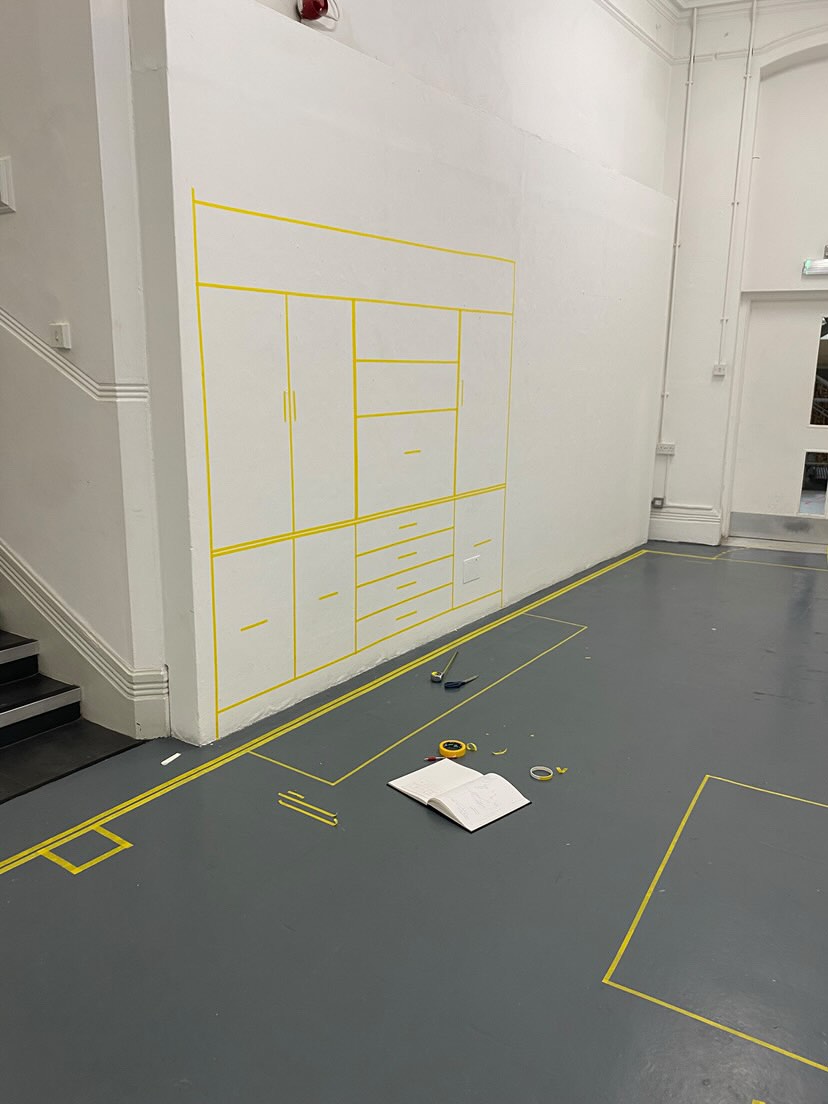 A wall with an outline of a wardrobe and drawers made from yellow tape. Art supplies and an open notebook are on the gray floor nearby. The scene is in a spacious, white-walled room with stairs to the left.