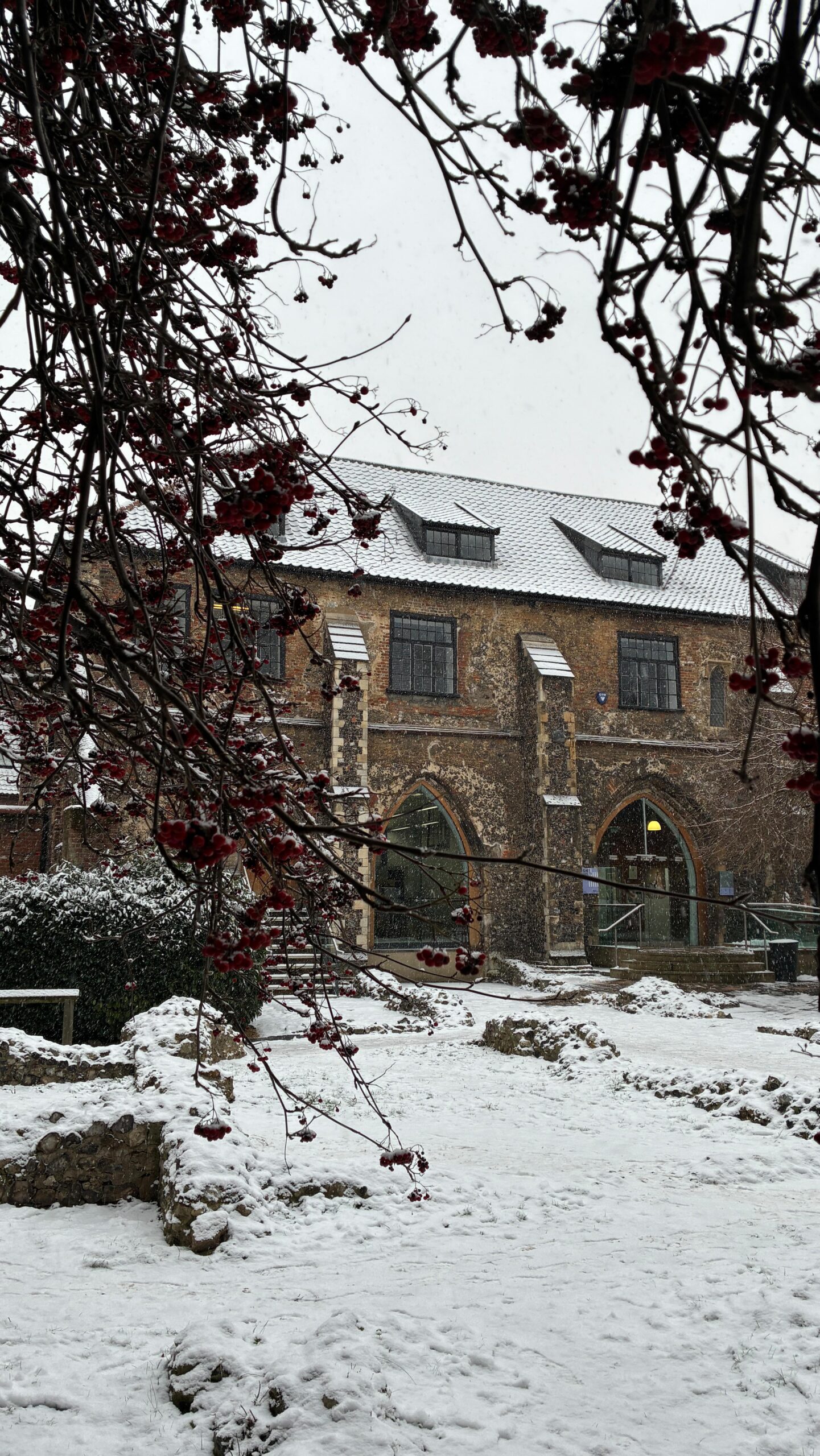 A historic brick building with arched windows and doors is covered in snow. Snow blankets the ground and branches with red berries in the foreground, creating a wintry scene.