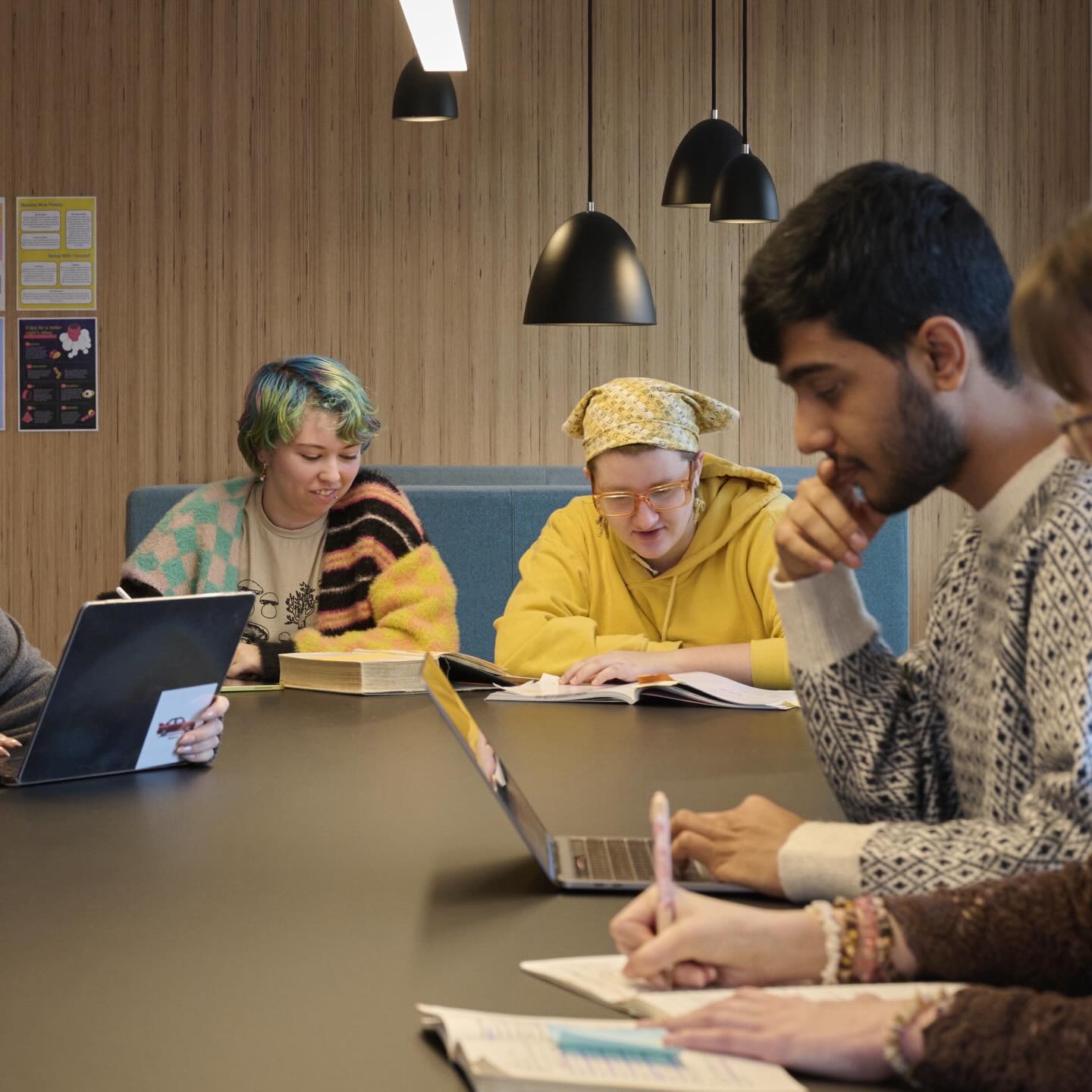 A group of five people sit at a table, studying and writing in notebooks. One person uses a laptop, another wears a yellow hoodie and headscarf, while another has colorful hair. Hanging lamps and a wood-paneled wall are in the background.