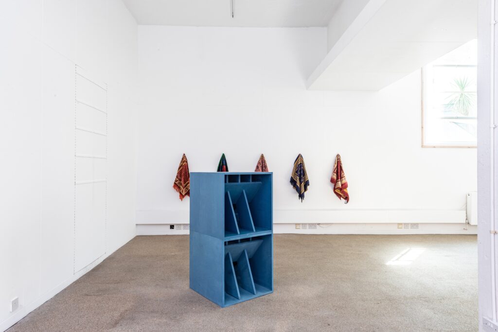 A minimalist gallery space with white walls and carpeted floor, featuring a blue geometric sculpture in the center and five colorful textiles hanging on the wall in the background. Light streams in from a high window.