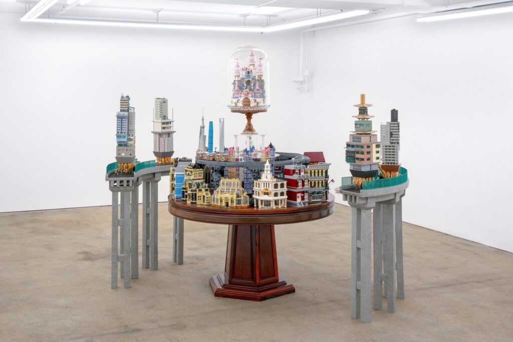 A round wooden table in the east gallery displays a detailed miniature city with colorful buildings and towers. The city extends onto raised platforms supported by gray pillars, all set in a white-walled space.