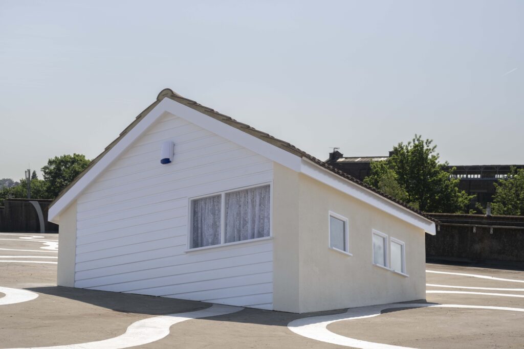 A small white house with closed windows and lace curtains appears partially submerged into a flat, paved surface, with trees and a building visible in the background under a clear sky.