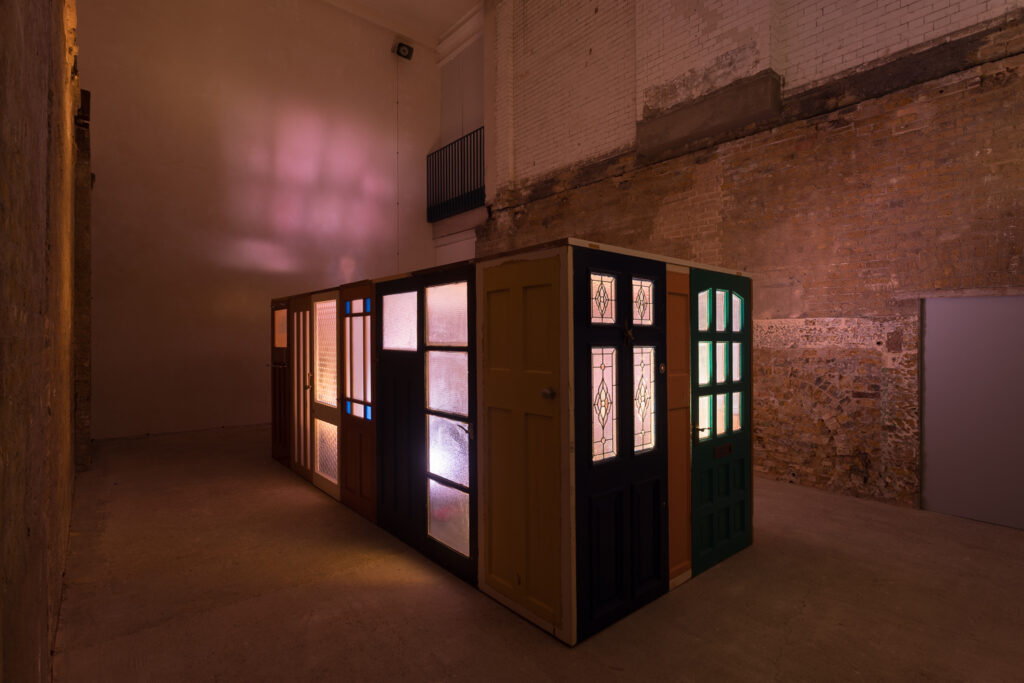 A dimly lit room features a large rectangular structure made of various old doors and frosted glass panels, illuminated from within by warm, colorful lights that cast shapes onto the surrounding brick walls.