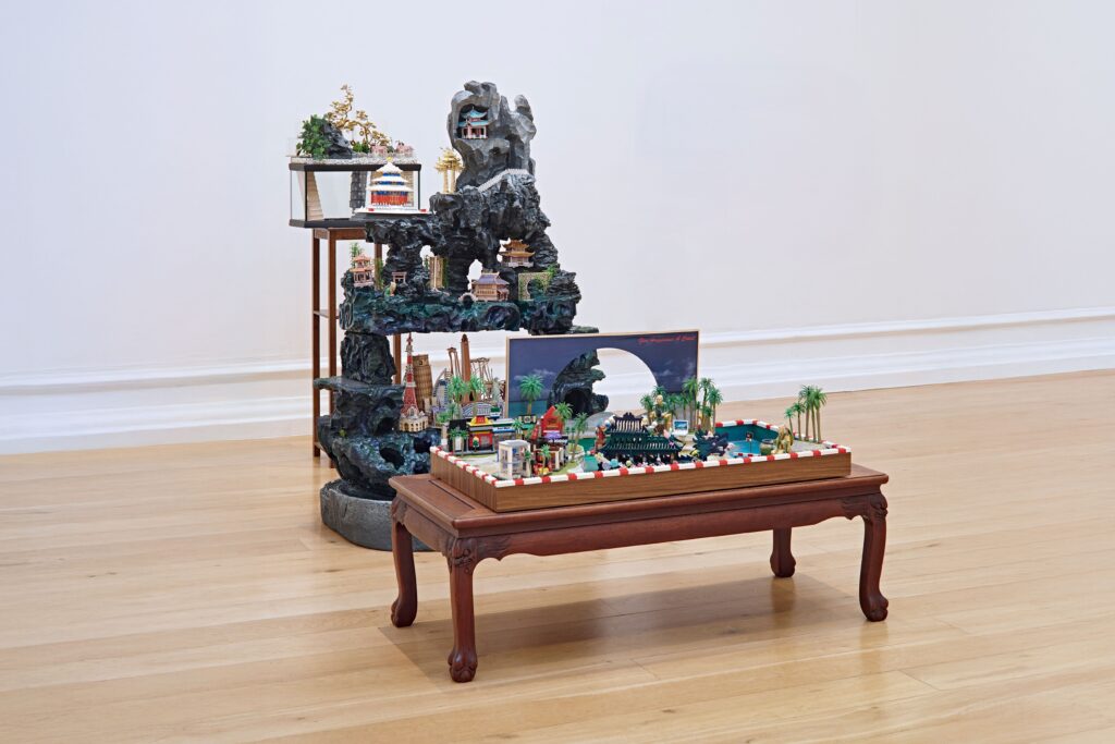 A small wooden table displays a detailed miniature landscape diorama with palm trees and buildings; behind it, another stand in the east gallery holds an artificial rock formation with tiny houses and plants, both set on a wooden floor.